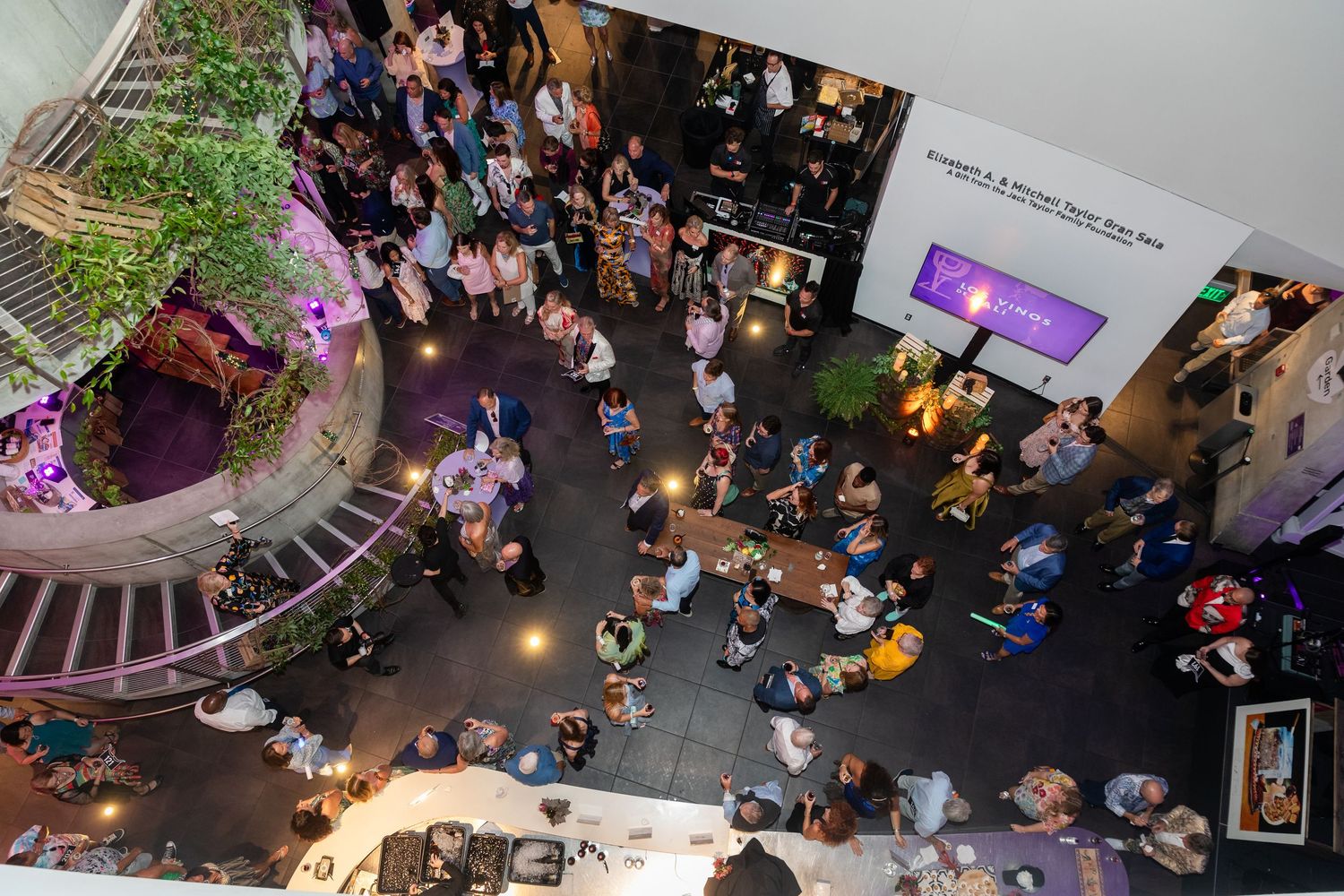 Aerial view of a crowded indoor event space with guests mingling beneath spiral stairs and purple accent lighting.