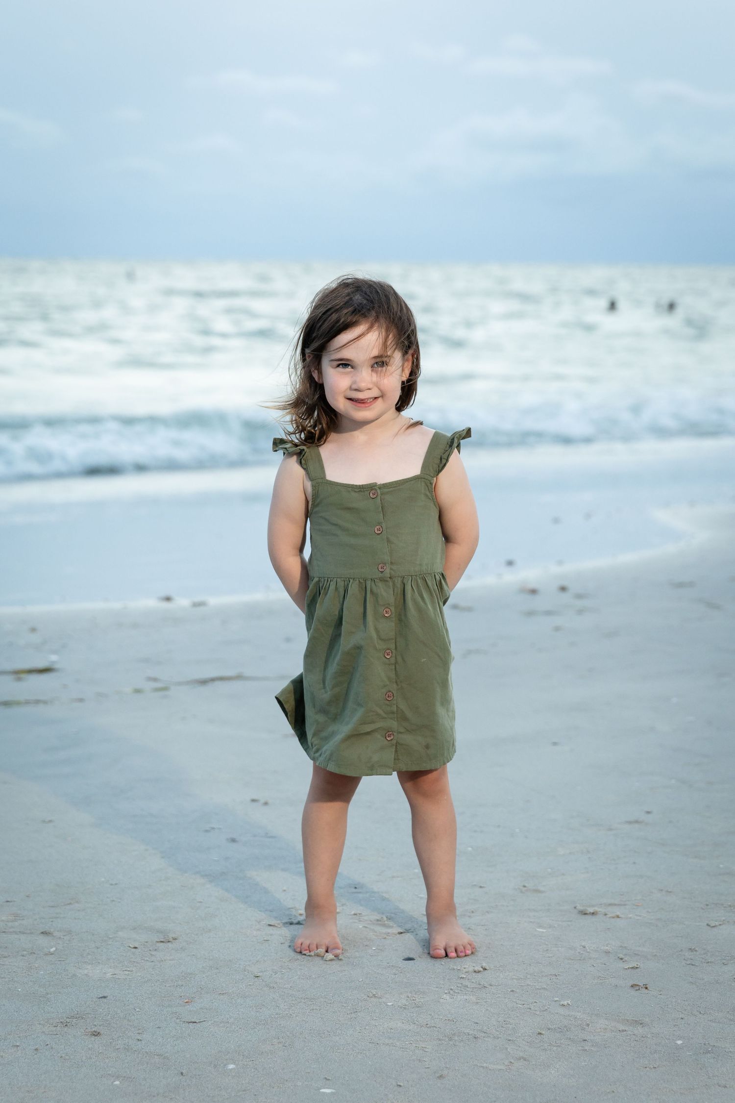 A young girl in a green dress stands barefoot on a sandy beach with ocean waves in the background.