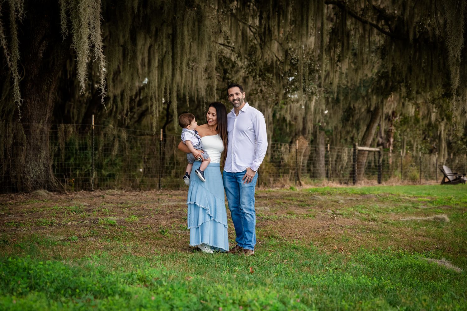 Family of three posing outdoors under Spanish moss trees on green grass.