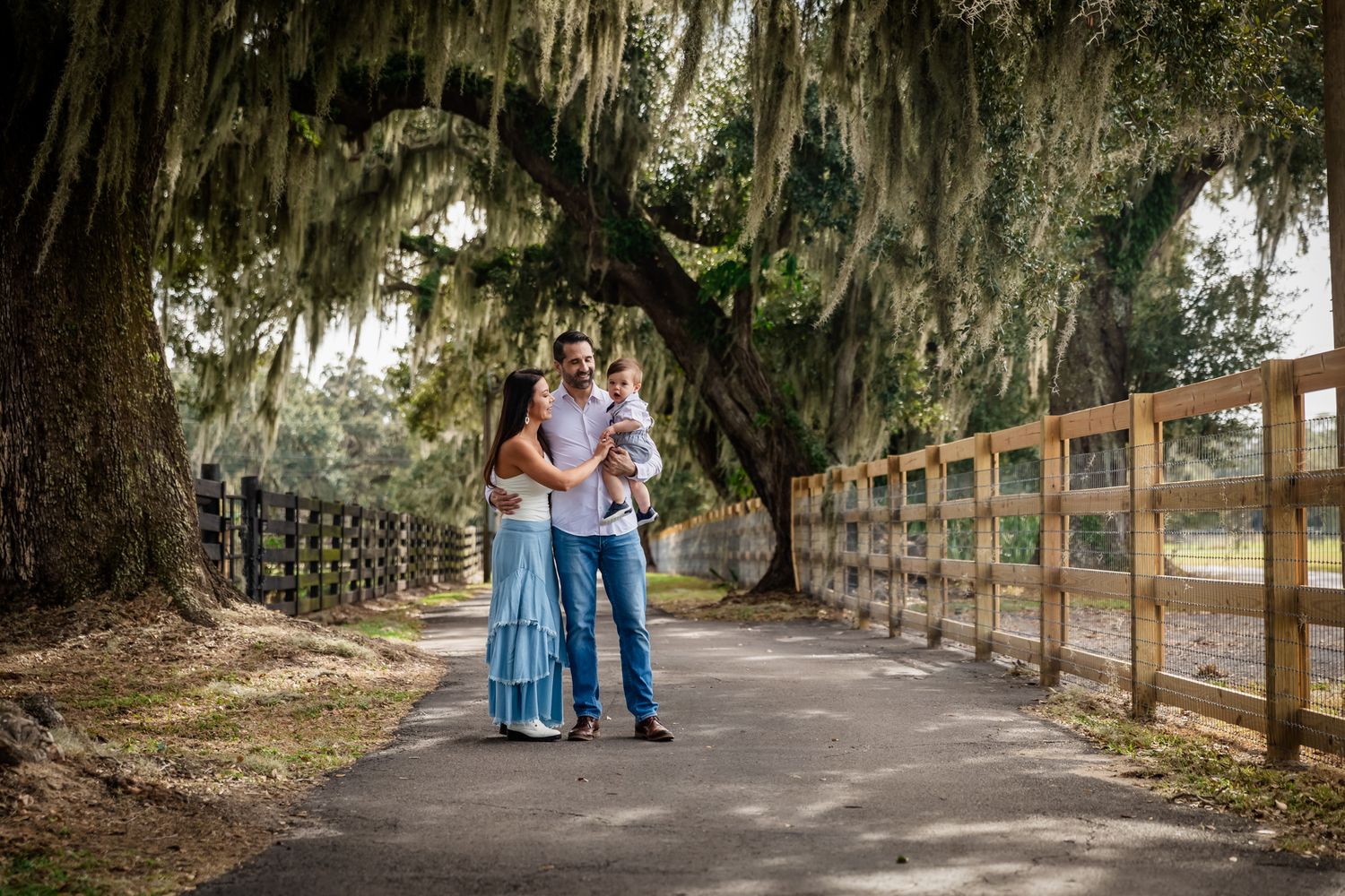 A couple stands on a tree-lined path draped with Spanish moss, holding their baby between them near a wooden fence.