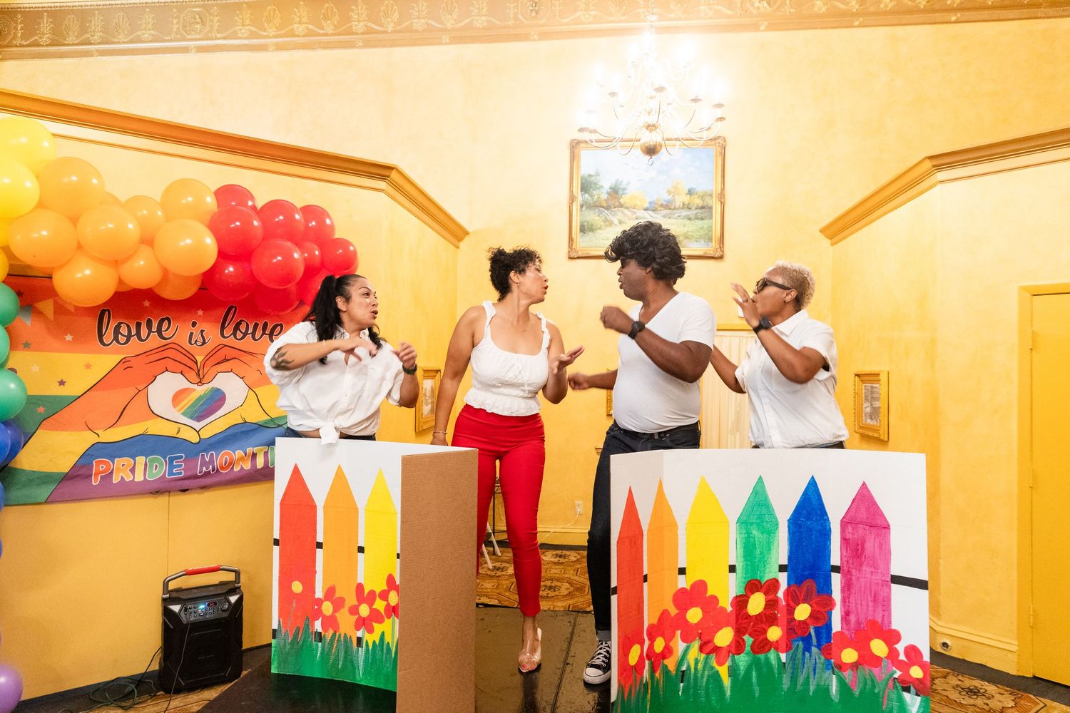Four people interact near colorful Pride-themed decorations with rainbow balloon arch backdrop.