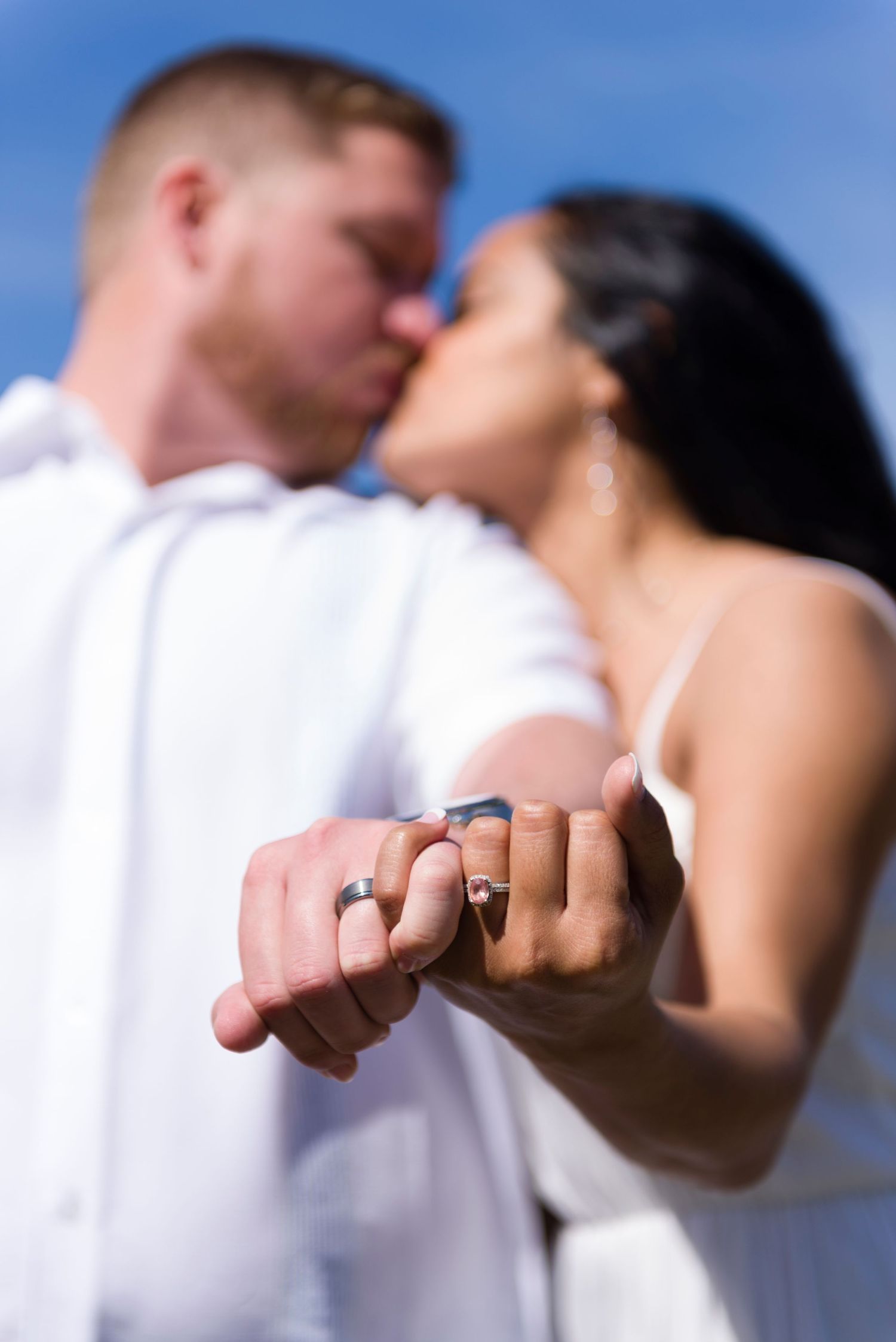 Newlywed couple kissing while holding hands displaying wedding rings against a blue sky backdrop.