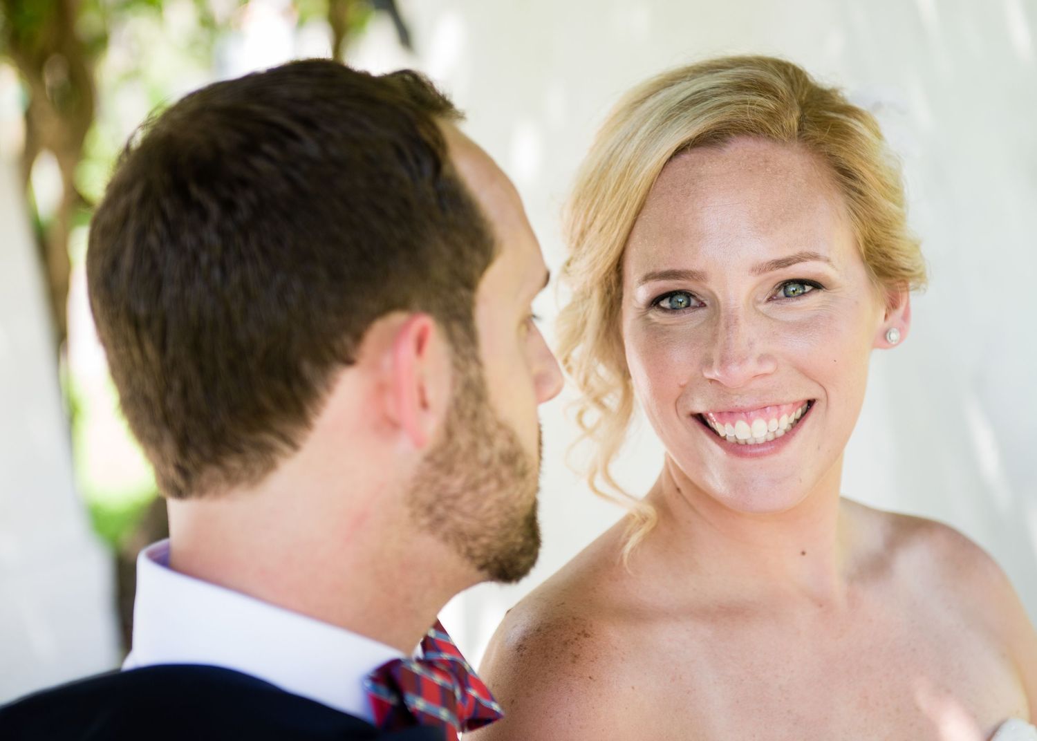 Smiling blonde bride with freckles gazes at groom in blue suit with red tie at outdoor wedding ceremony.