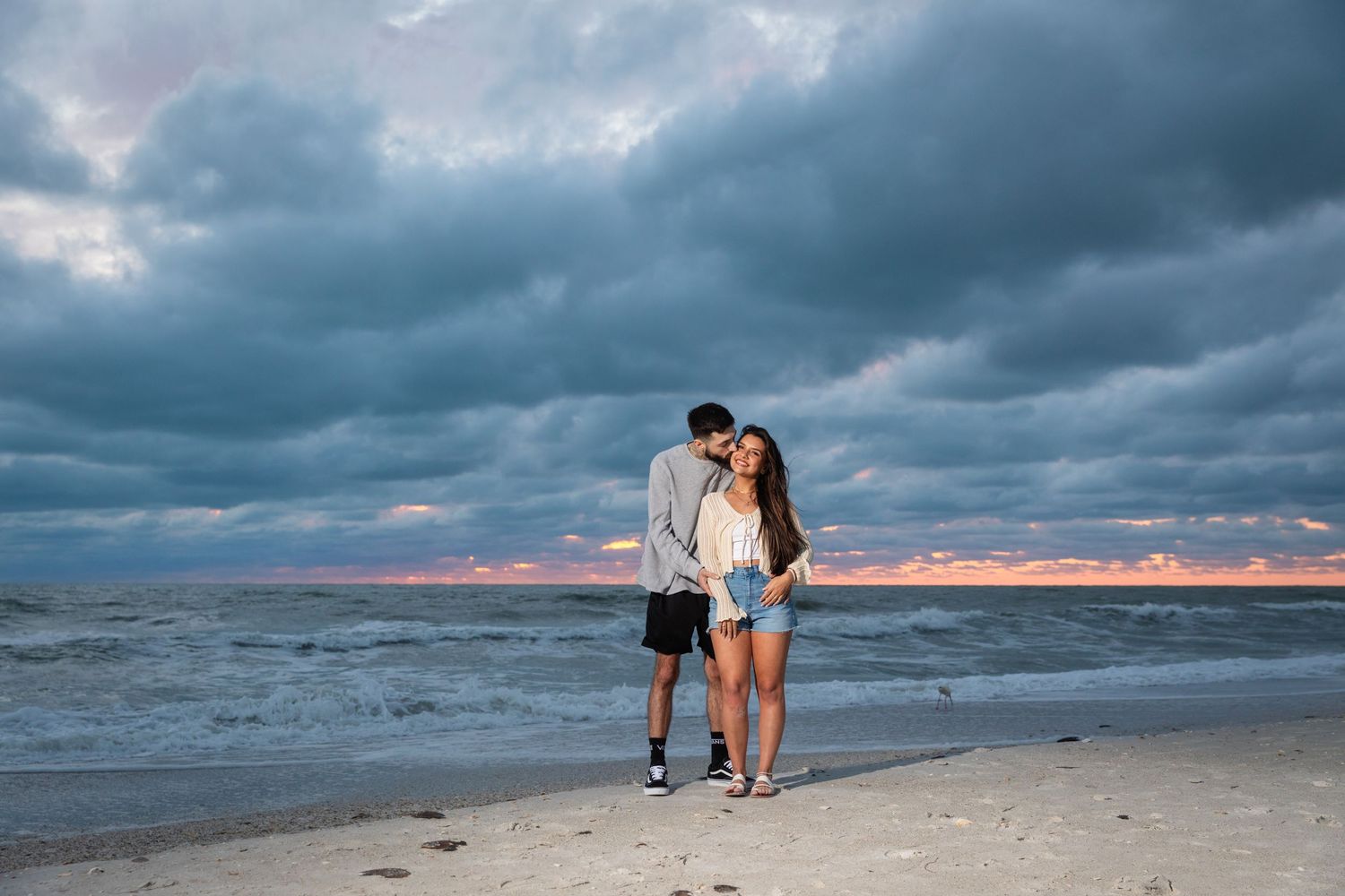 Couple embracing on a sandy beach at sunset with dramatic storm clouds over the ocean.