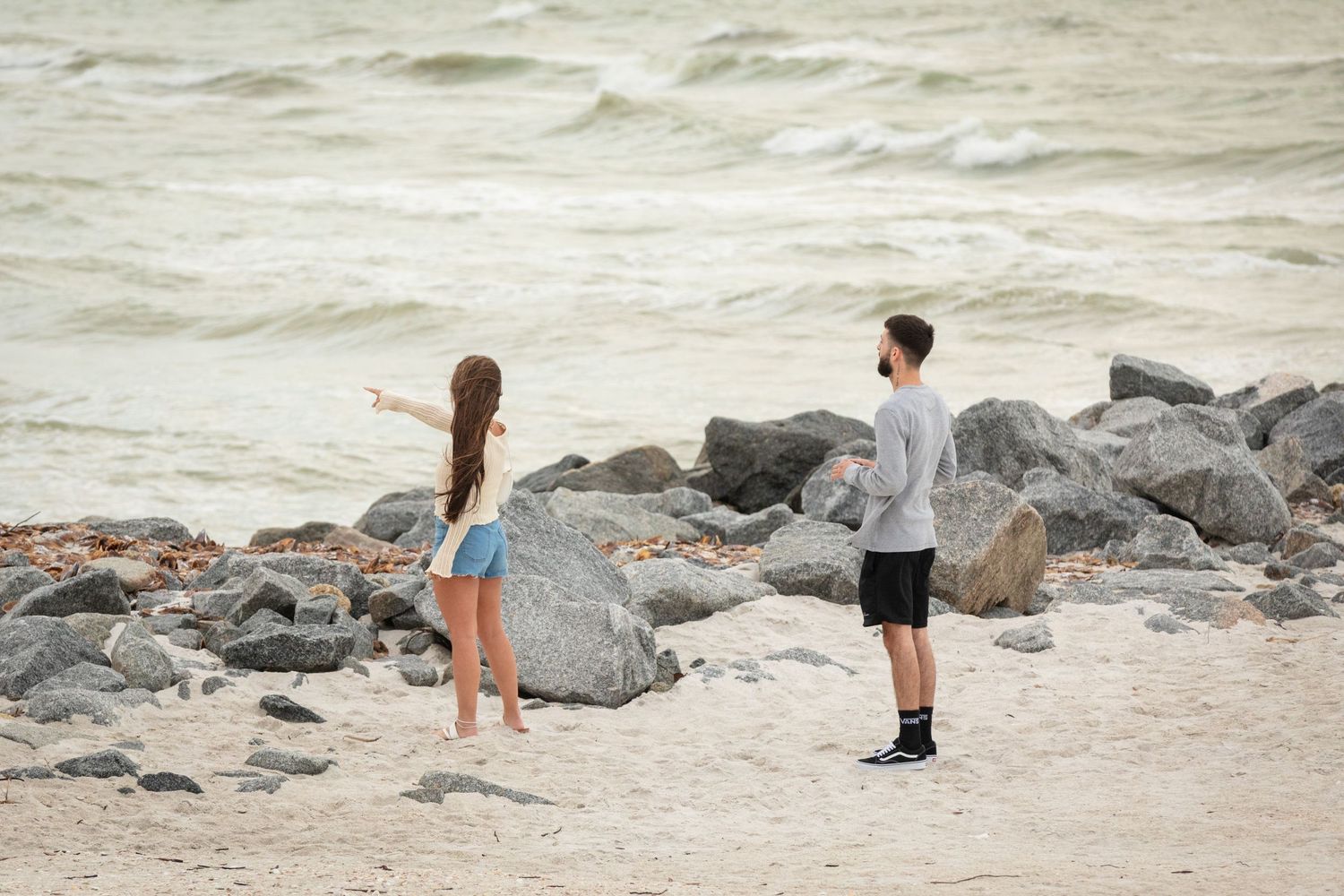 A girl and boy stand on a rocky beach, with the girl pointing toward the ocean waves.