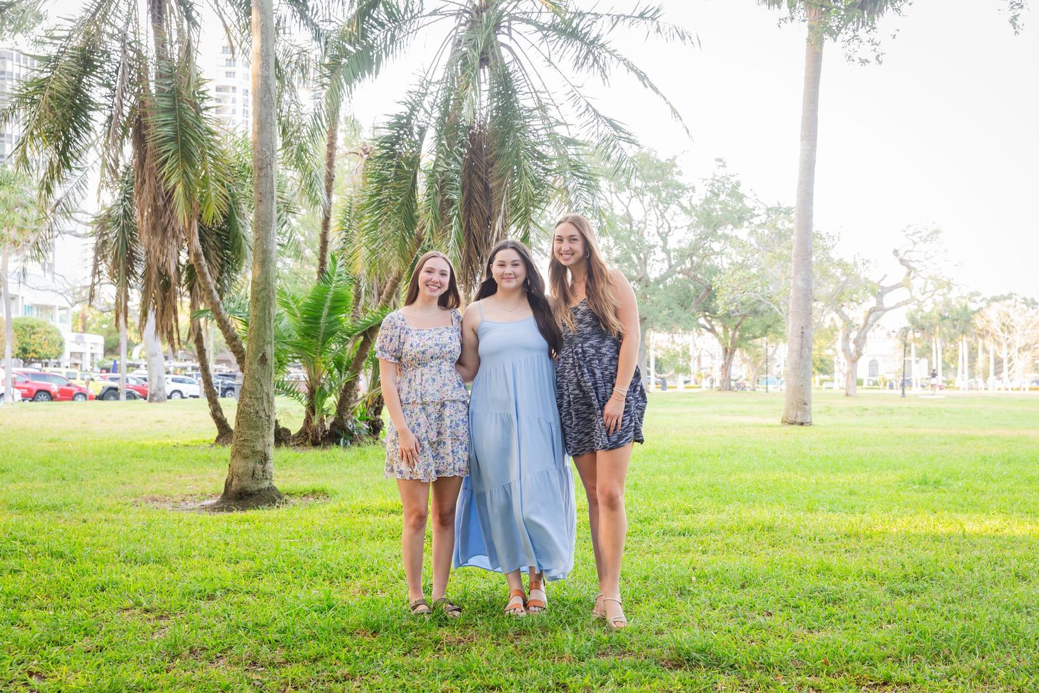 Three young women in summer dresses smiling together on green grass near tropical palm trees outdoors.