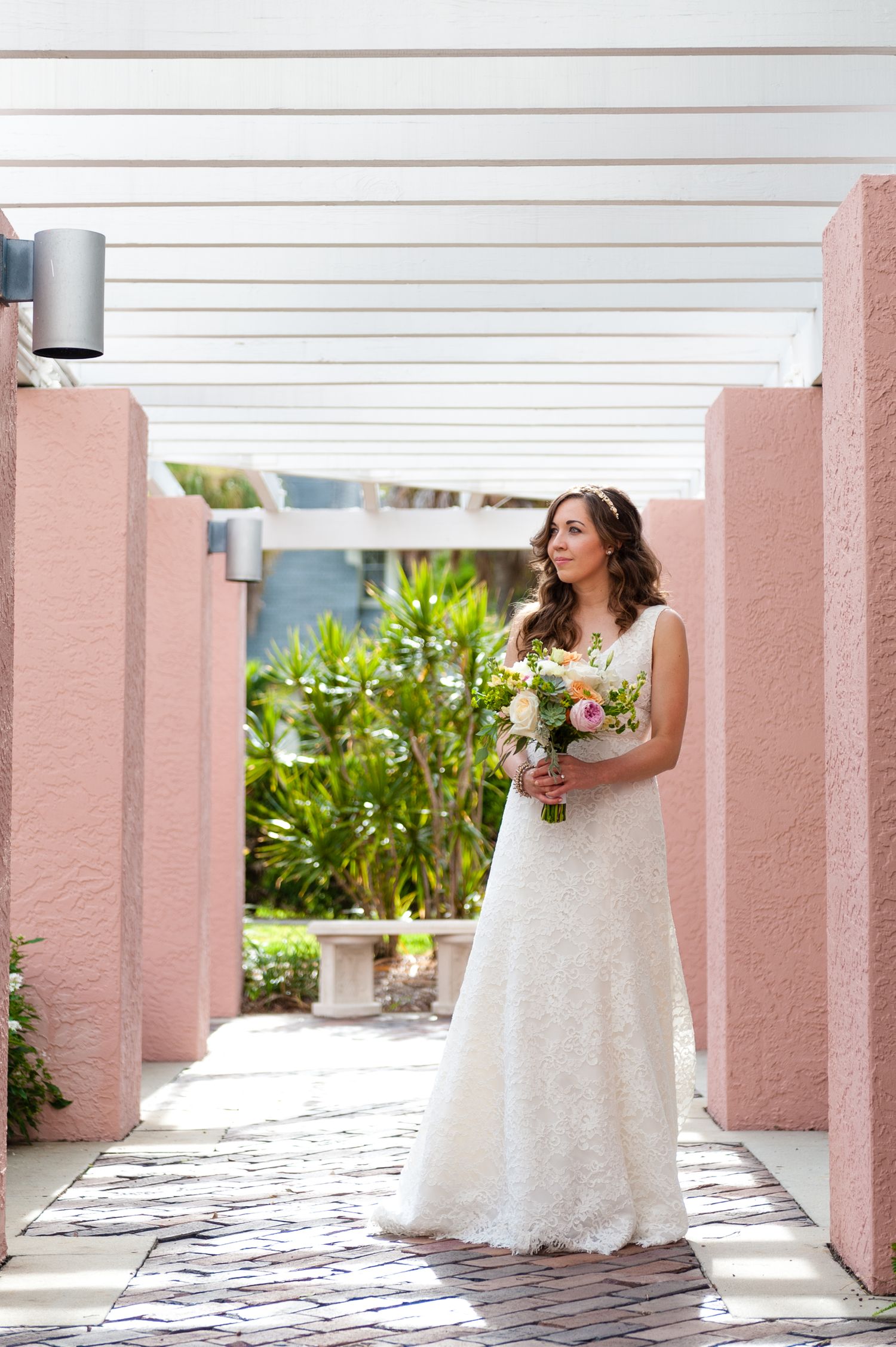 Bride in white lace gown holding bouquet between pink columns at outdoor venue.