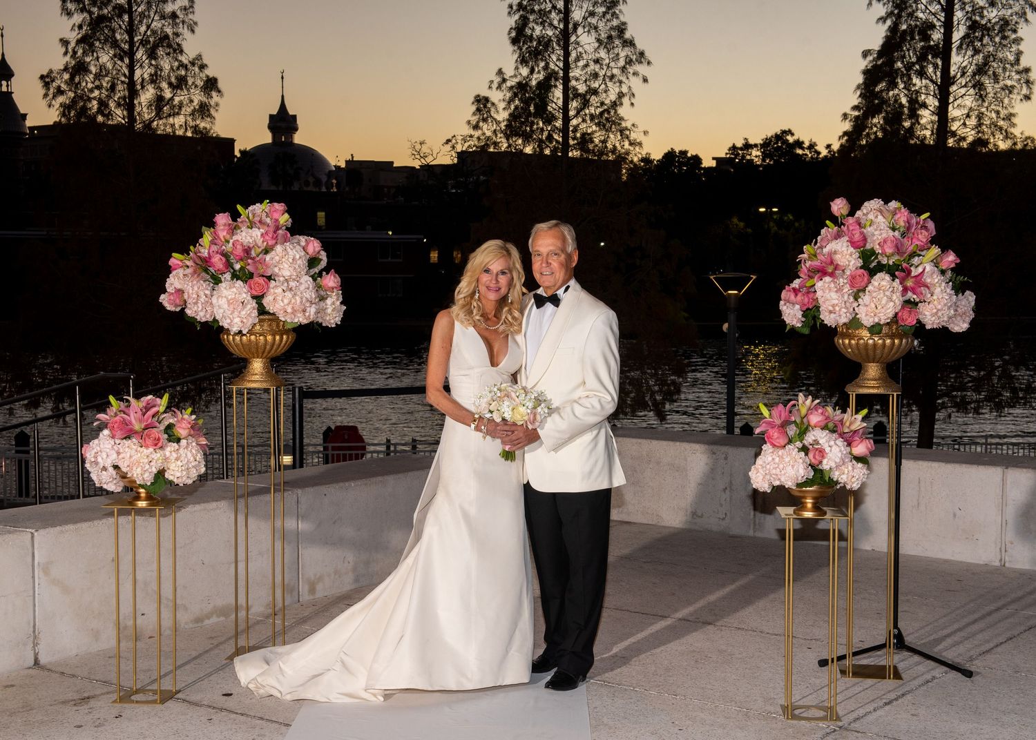 Bride and groom pose at elegant outdoor wedding ceremony with pink floral arrangements and waterfront backdrop at dusk.