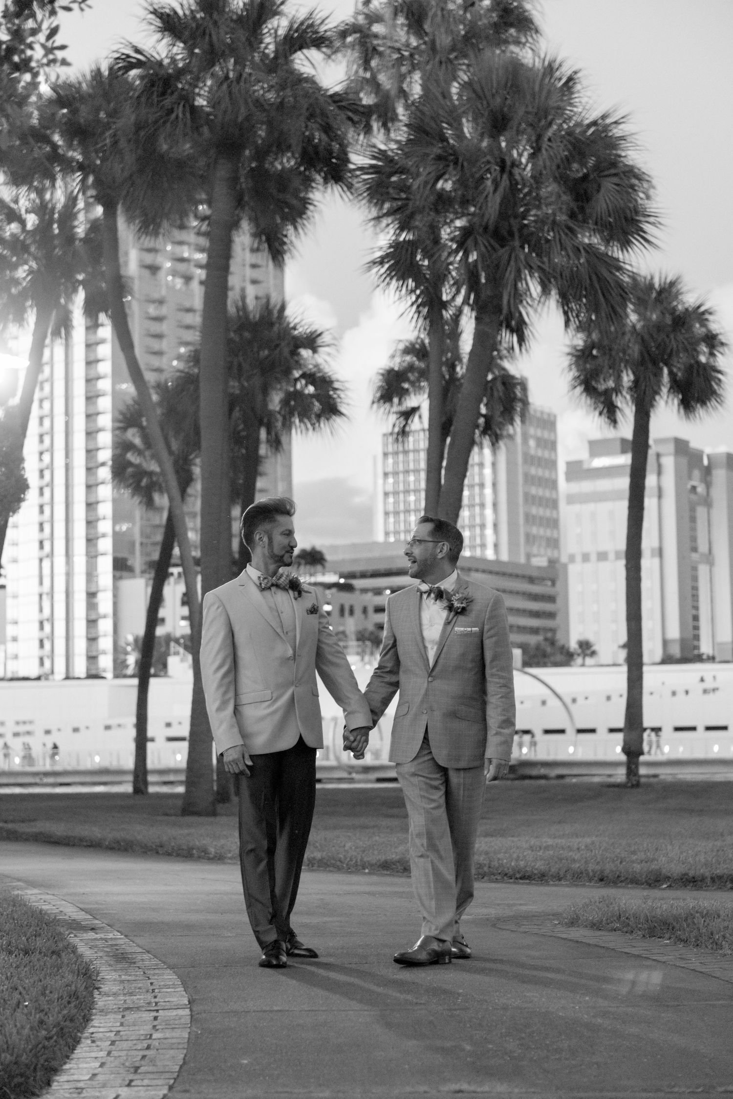 Two grooms hold hands and smile at each other on a palm-lined path with a city skyline backdrop.