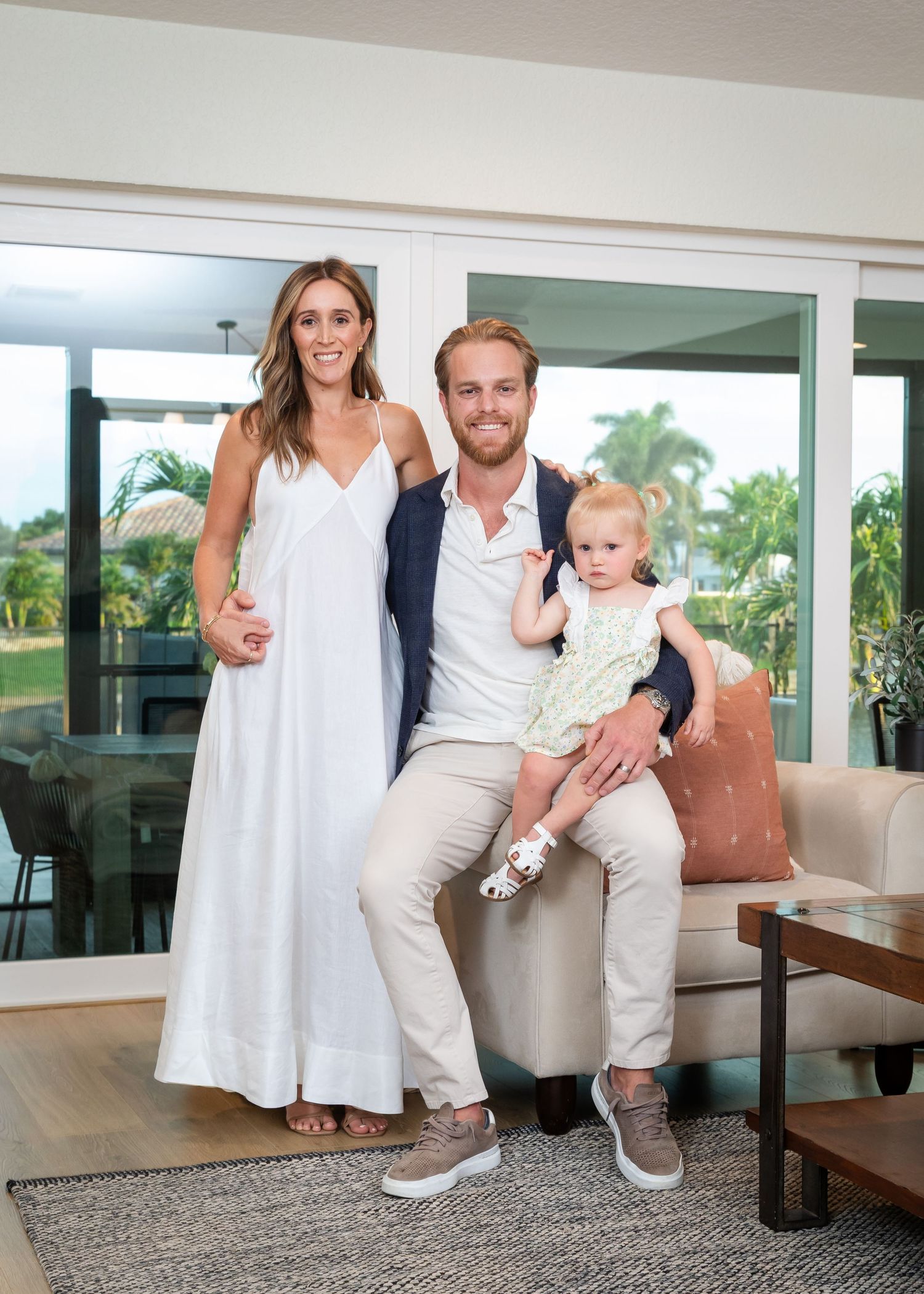 A happy family of three poses indoors, with a woman in white dress, man in navy blazer, and baby girl.