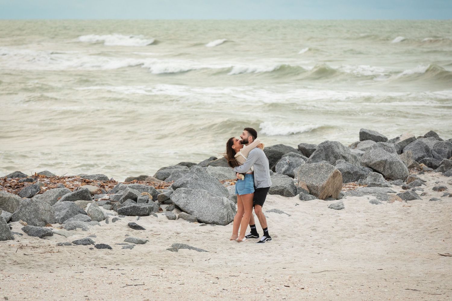 Couple embracing on a sandy beach with rocky shoreline and ocean waves in the background.