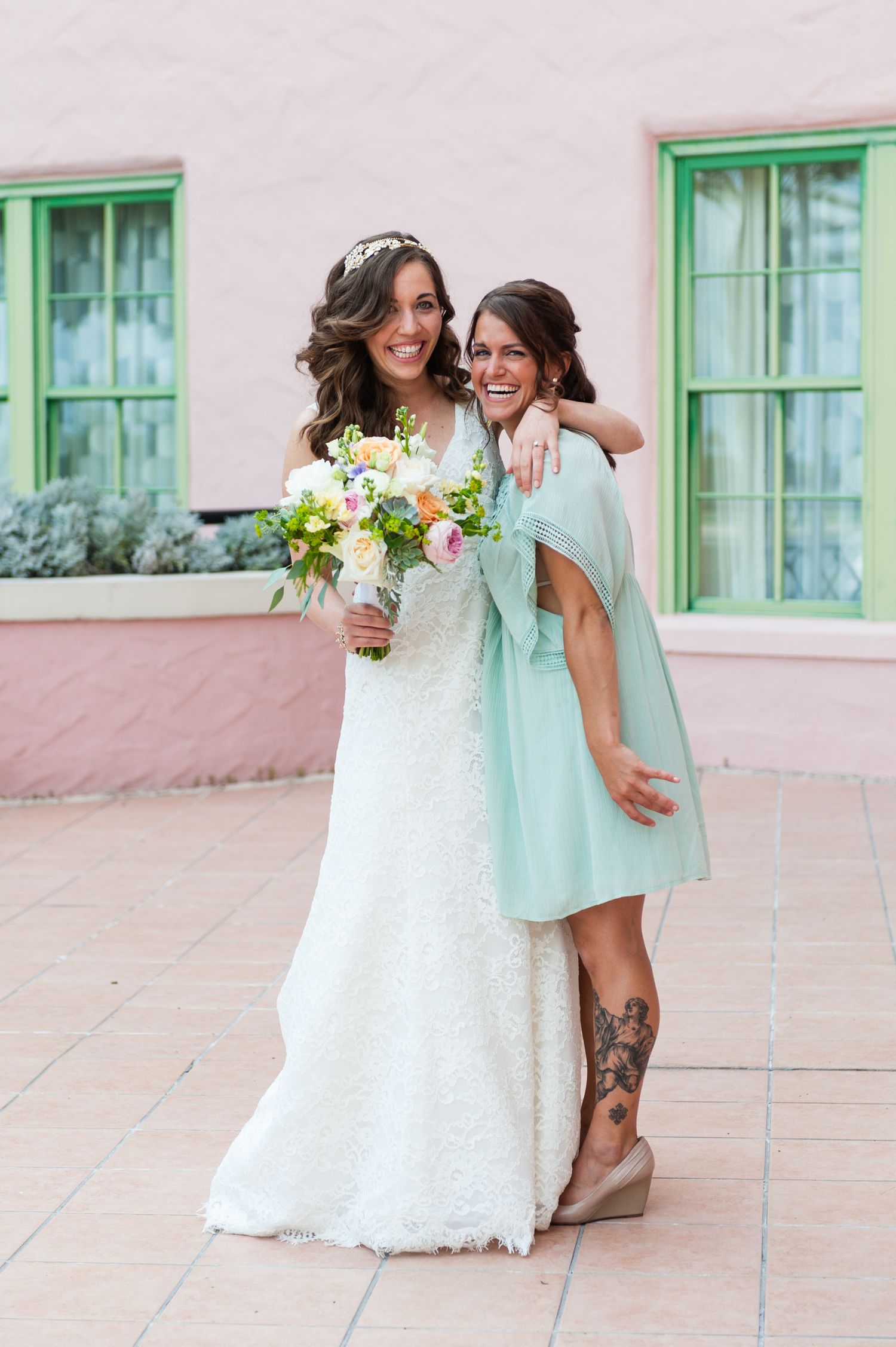 Bride in white lace gown hugging bridesmaid in mint dress while holding floral bouquet outdoors at The Vinoy Resort.