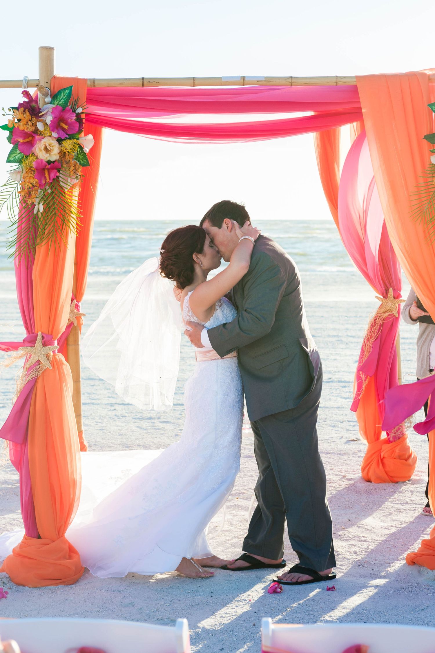 Bride and groom share first kiss under vibrant orange and pink draped arch at beach wedding ceremony.