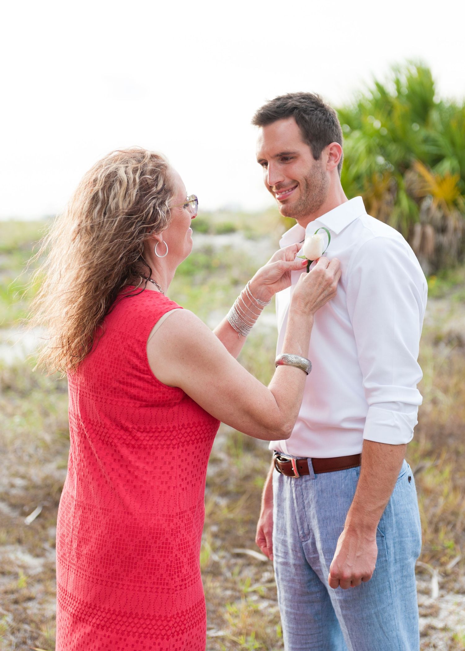Mother pins boutonniere on smiling groom in white shirt at outdoor beach wedding preparation.