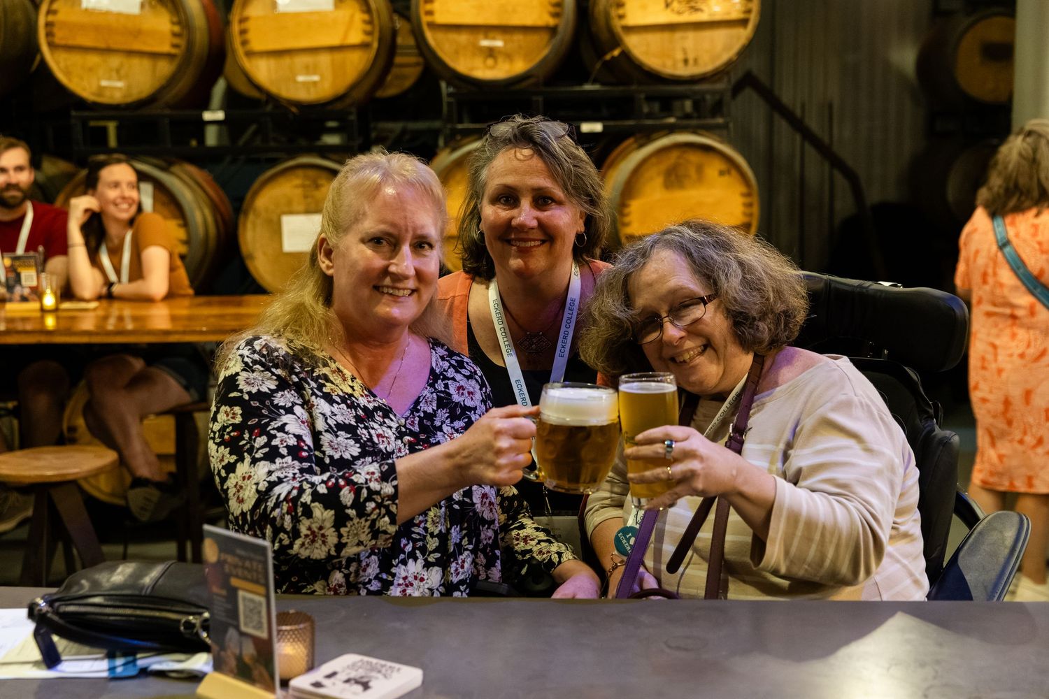 Three women toasting with beer glasses inside a brewery with wooden barrels in background.