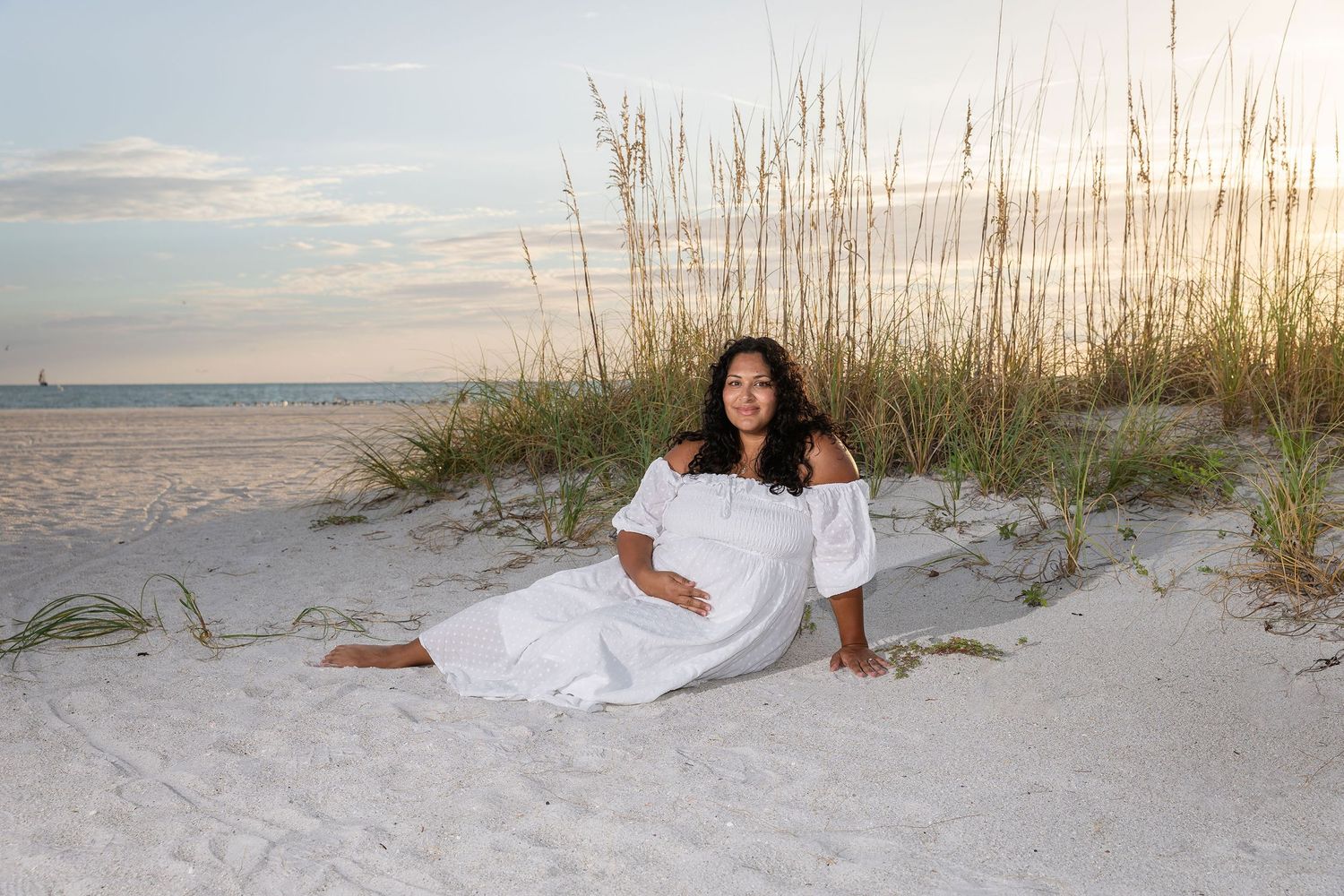 Pregnant woman in white dress sitting on white sand beach dunes at sunset with sea oats in background.