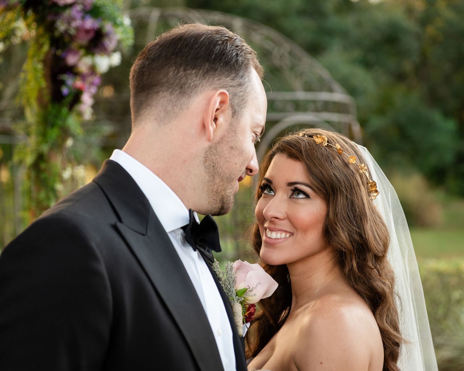 A bride and groom share a romantic moment outdoors, with the bride smiling in a veil and floral crown.