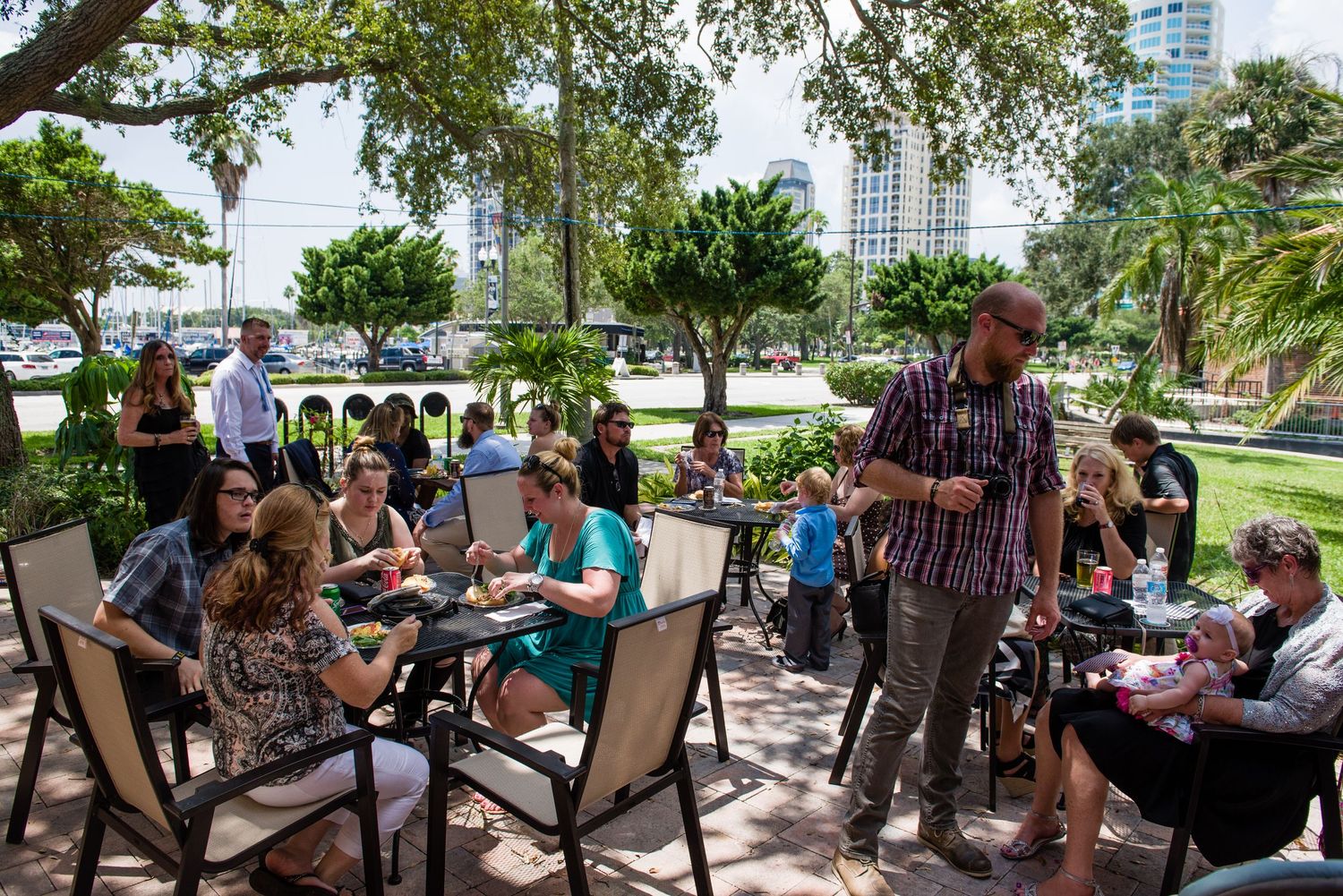 Group of people dining outdoors at a patio restaurant surrounded by lush greenery and city buildings on a sunny day.