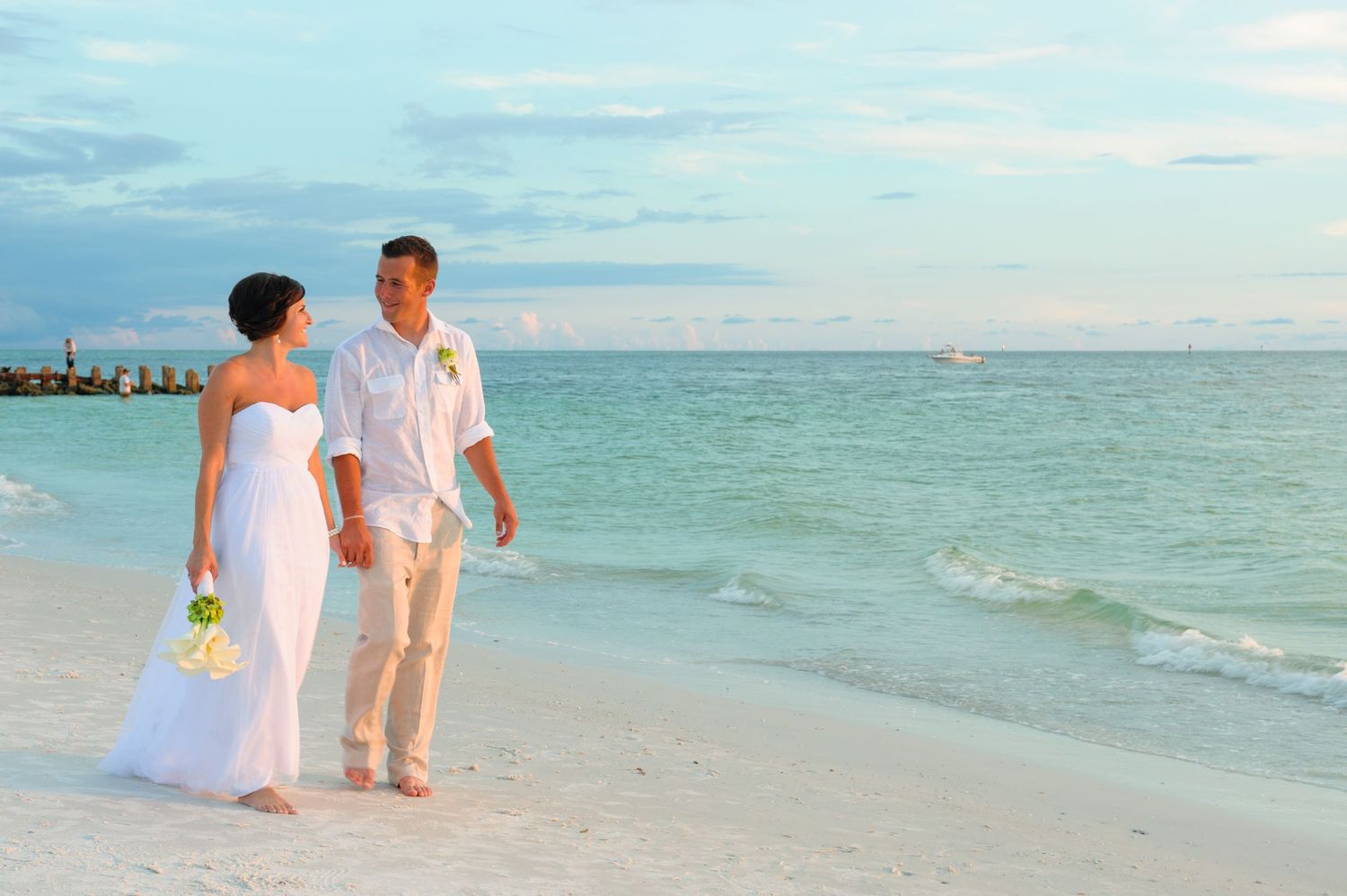 Bride and groom holding hands on a white sand beach with turquoise ocean waves during a romantic seaside wedding.