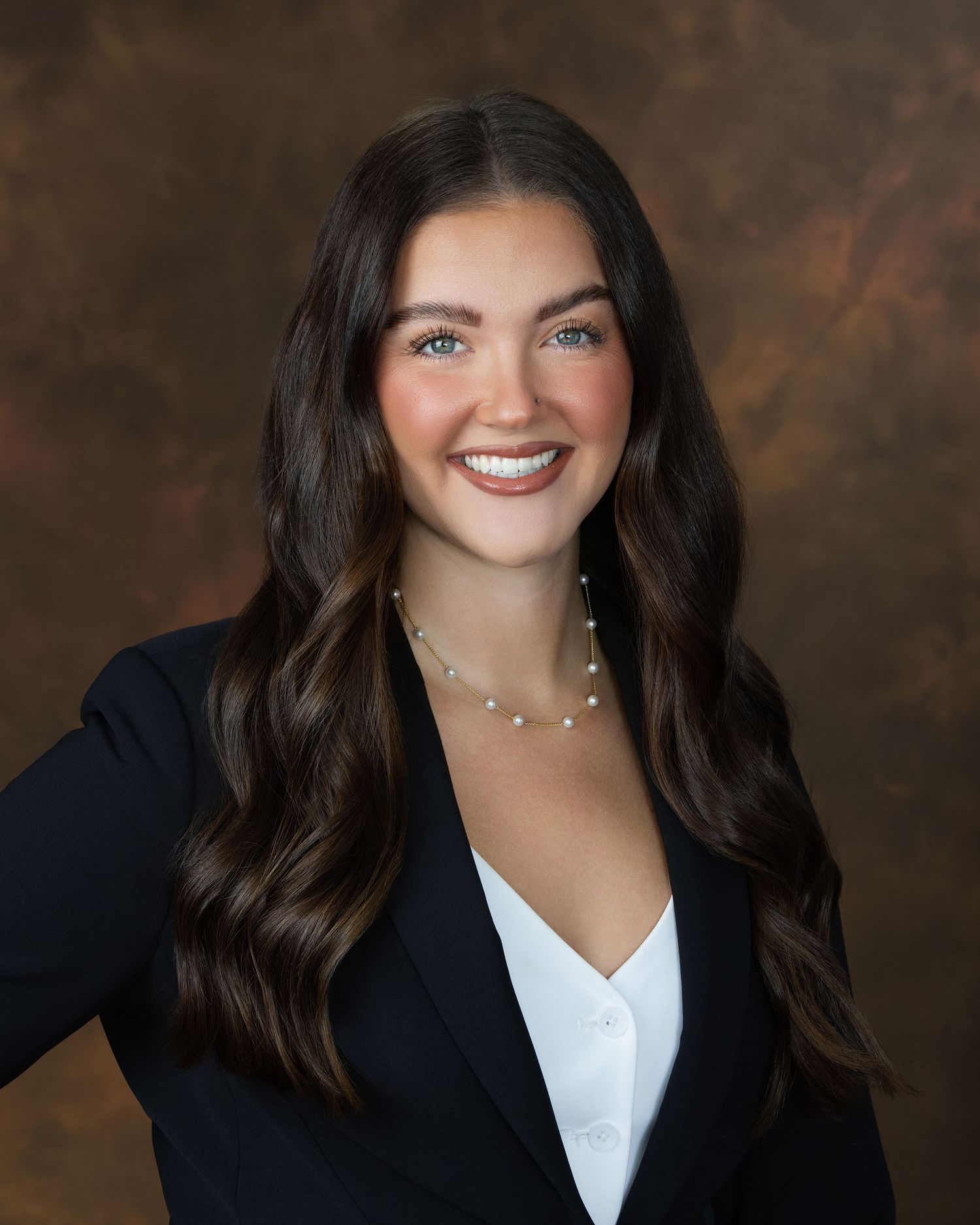 Professional headshot of a smiling young woman with long dark hair wearing a black blazer and pearl necklace.