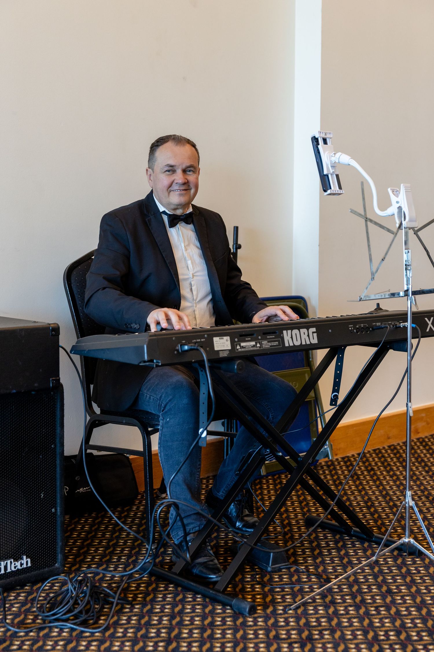 Man in tuxedo playing a Korg keyboard synthesizer while seated at an indoor event or wedding reception.
