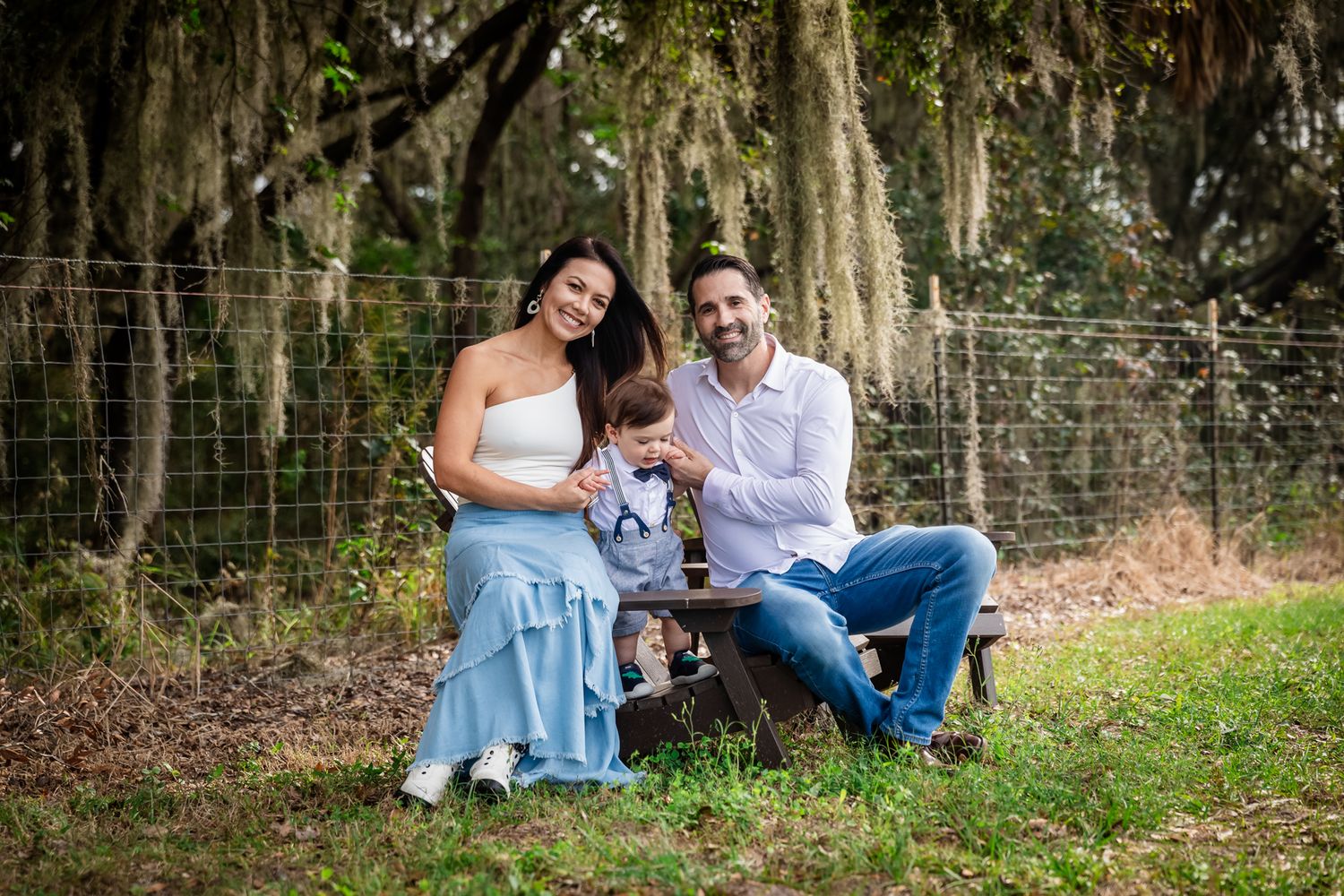 Happy family of three posing outdoors near moss-draped trees, parents smiling with toddler son in a rustic garden setting.
