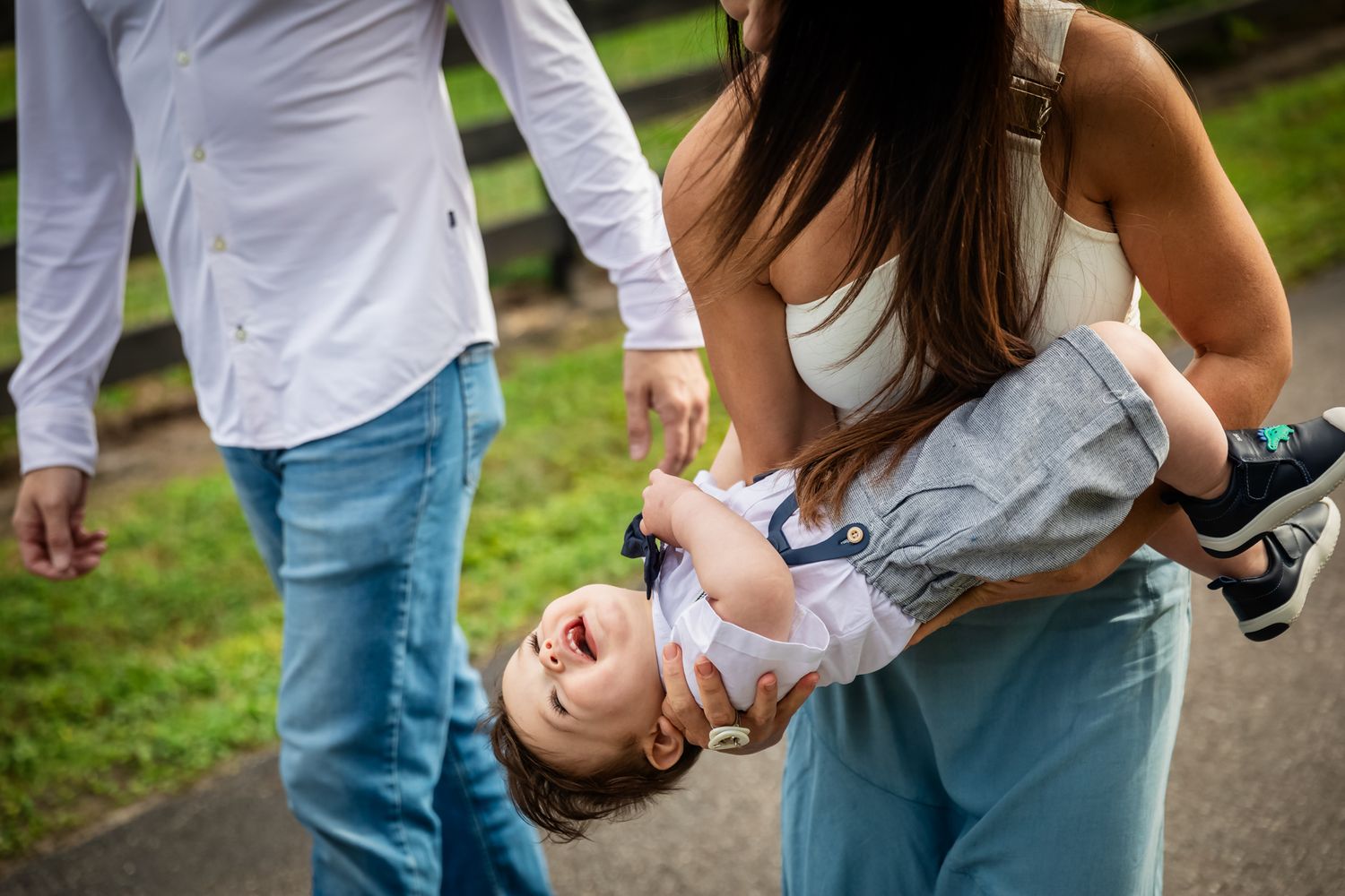 Parents playfully holding a laughing baby upside down outdoors on a paved path with green grass background.