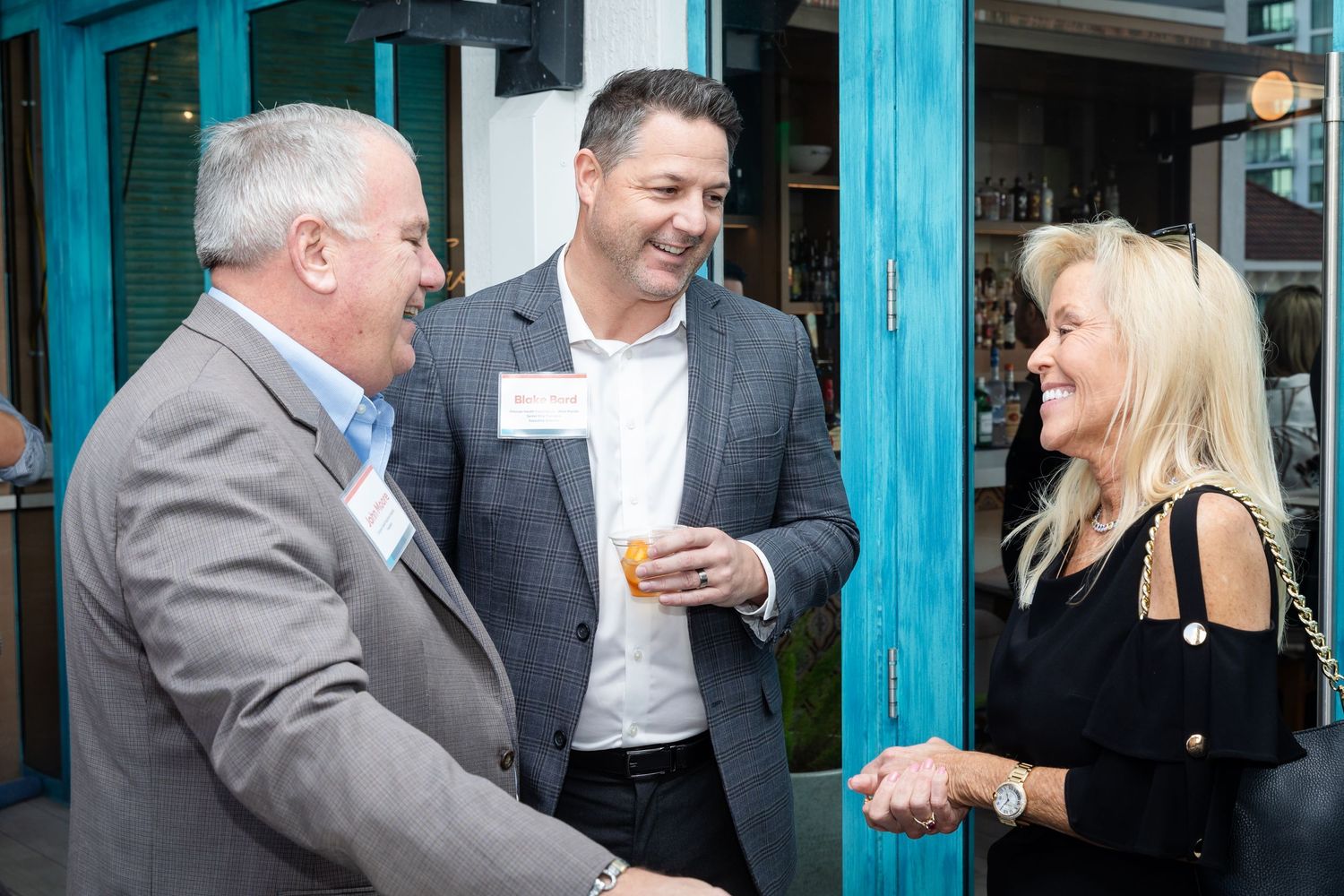 Three professionals networking and smiling at an outdoor business event, two men and a woman in business attire.