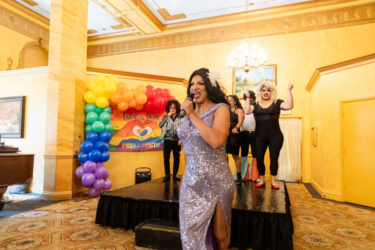 Drag queen in silver gown performs energetically before rainbow balloon display at Pride event.