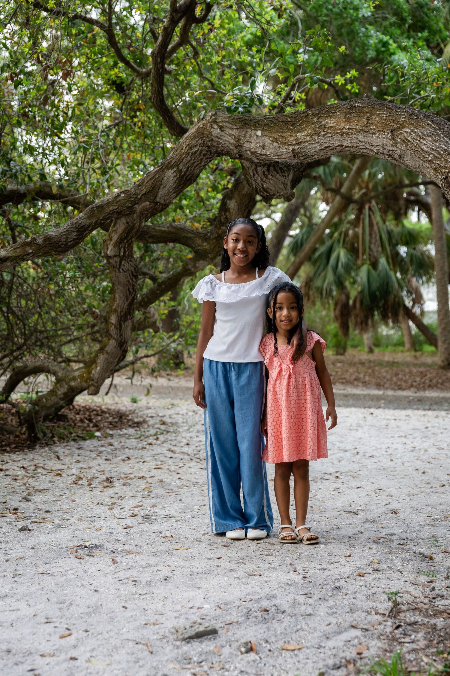 Two girls, one teenage and one young child, stand together under a large oak tree on a sandy path.