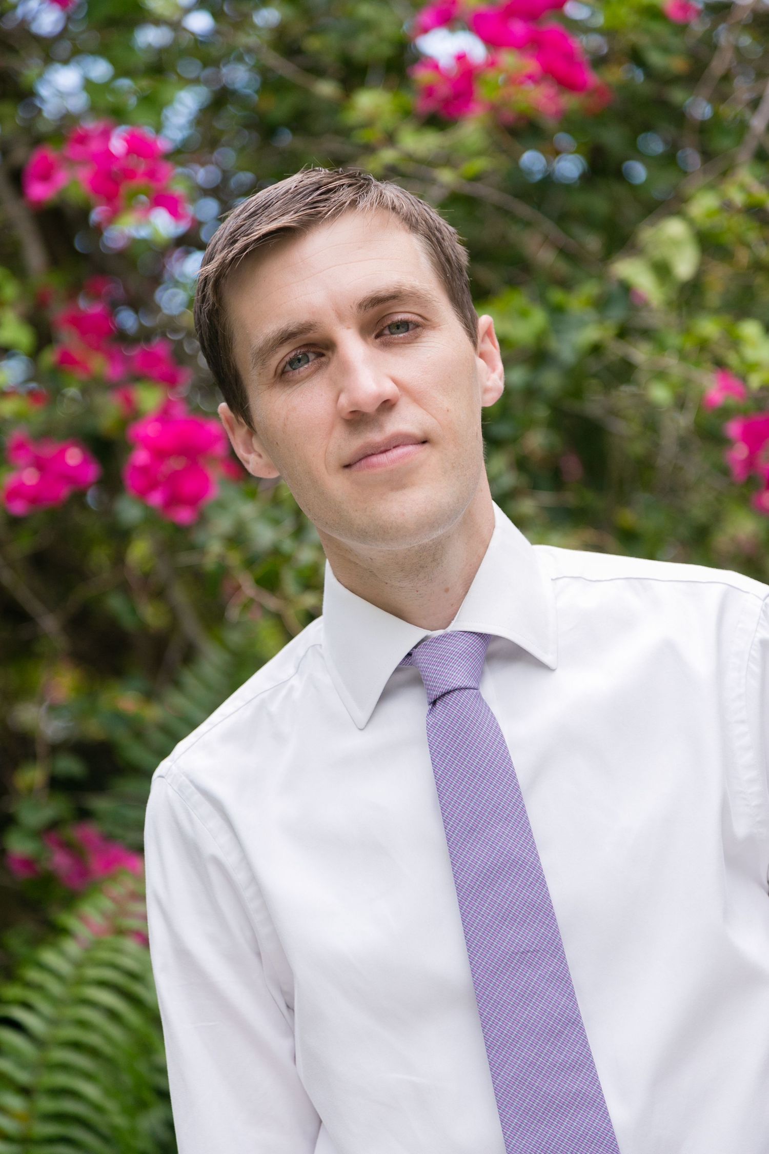 Young man in white dress shirt and lavender tie posing outdoors near pink flowers.