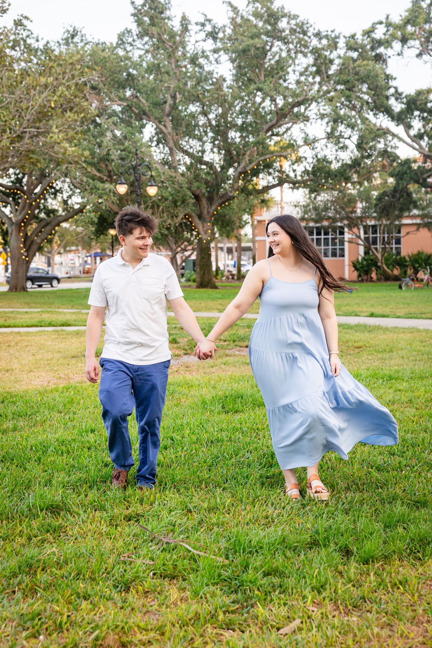 A couple holds hands and walks on green grass, the woman in a flowing light blue dress and the man in a white shirt and blue pants.