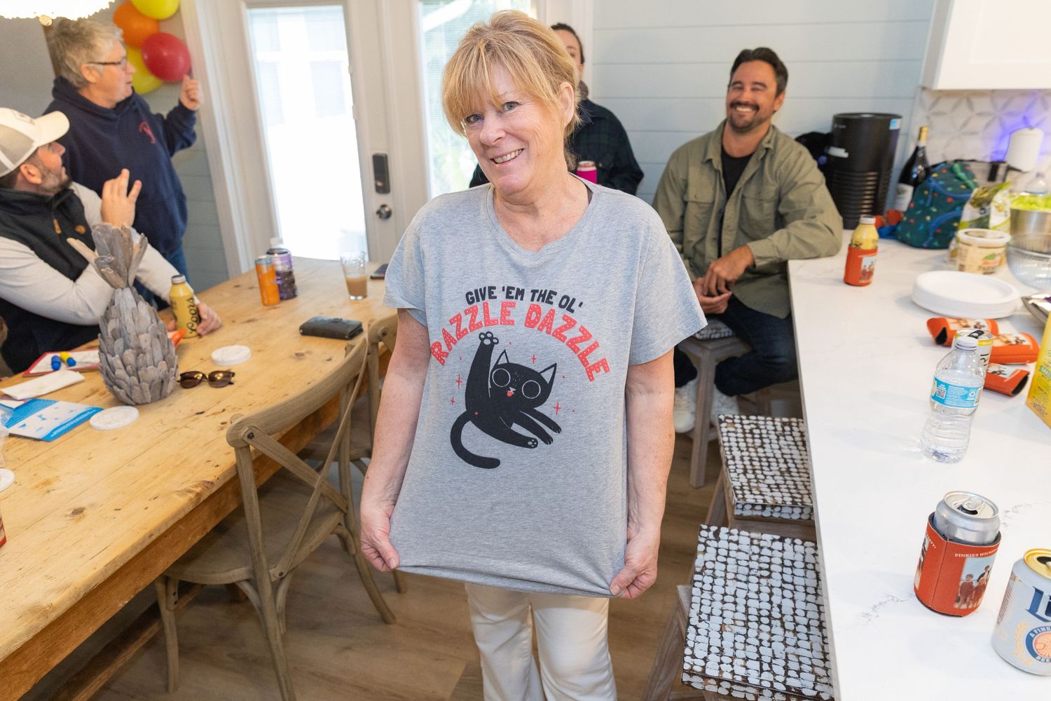 Woman smiling and holding up a gray cat graphic t-shirt at a casual indoor gathering with friends.