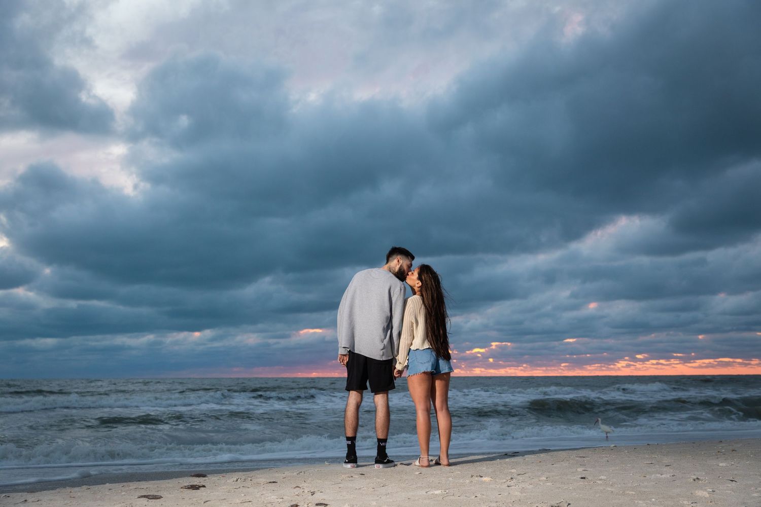 Romantic couple standing together on beach at dramatic sunset with stormy cloudy sky overhead.