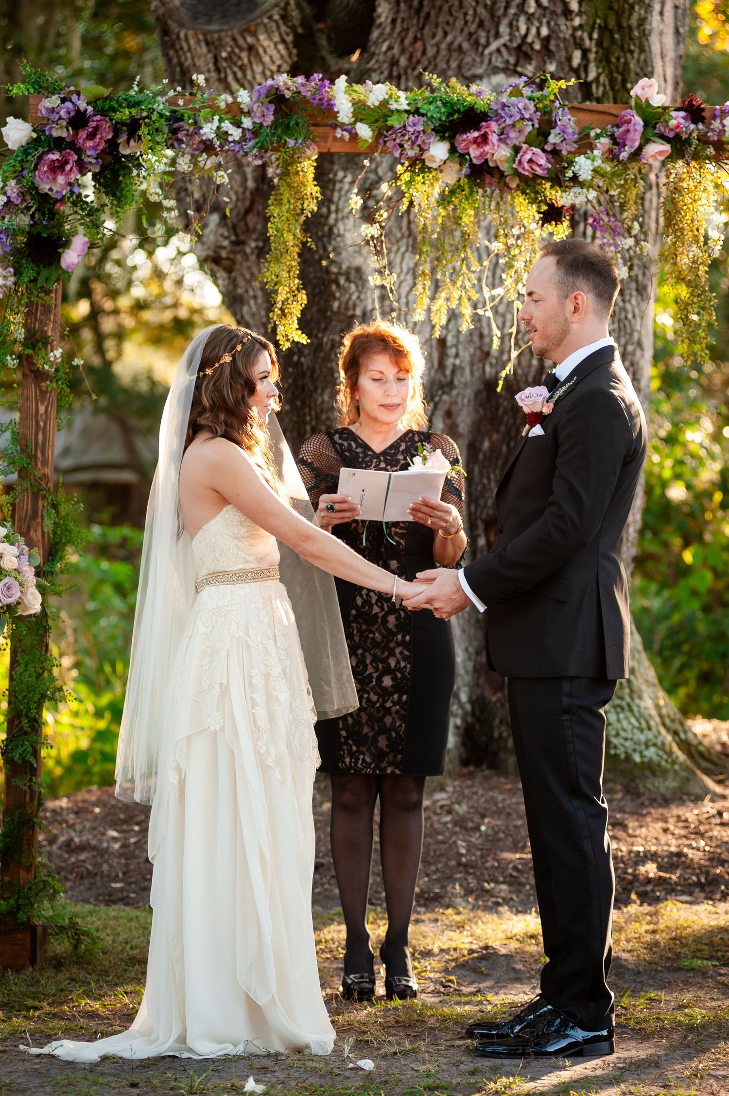 Bride and groom hold hands during outdoor wedding ceremony under floral arch with purple and pink blooms.