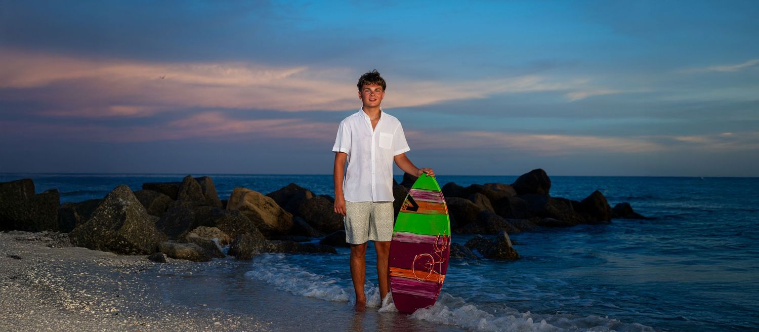 Young man holding a colorful surfboard on a rocky beach at sunset with vibrant pink and blue sky.