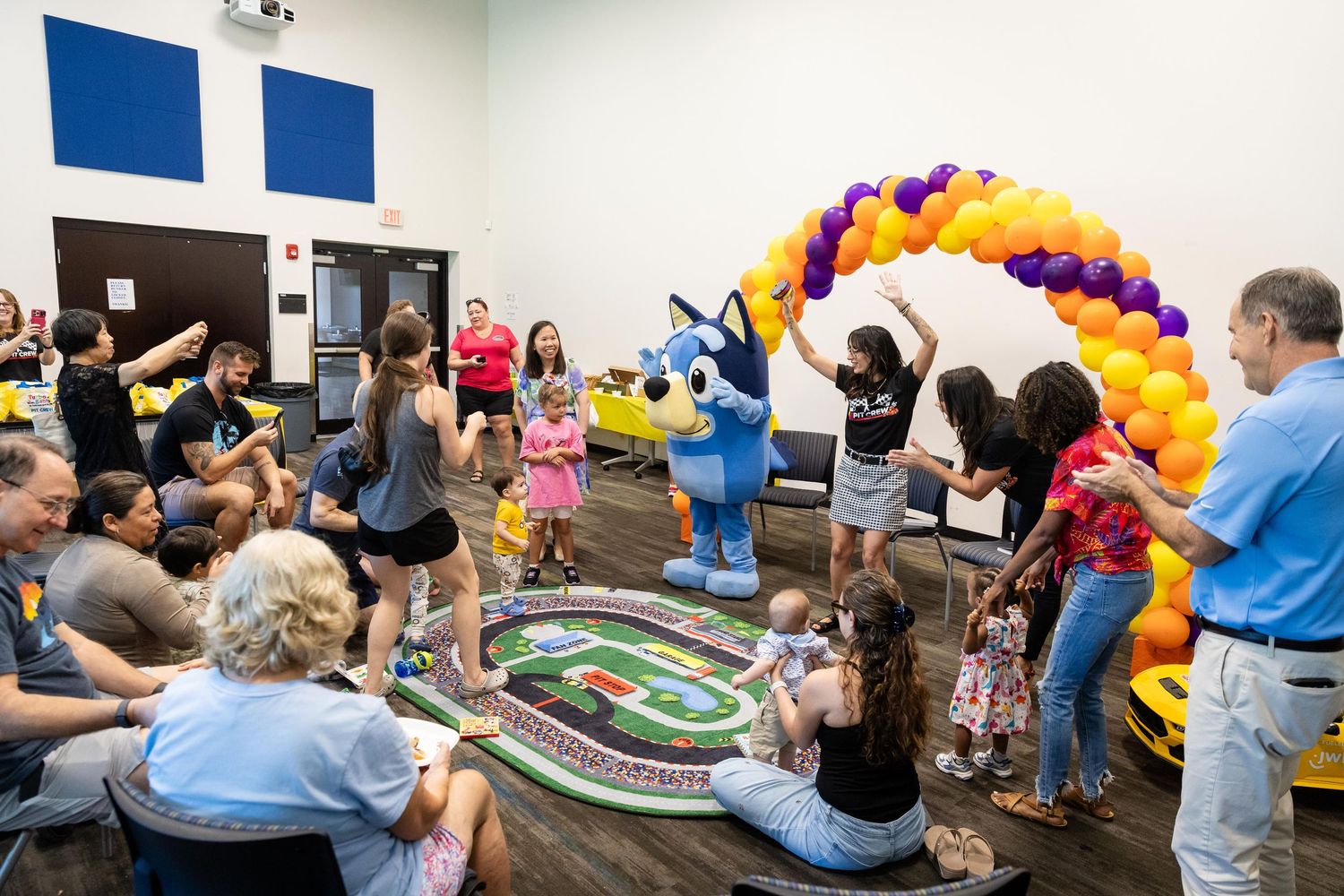 Children and families dance with a Husky mascot under a colorful balloon arch at a festive indoor event.