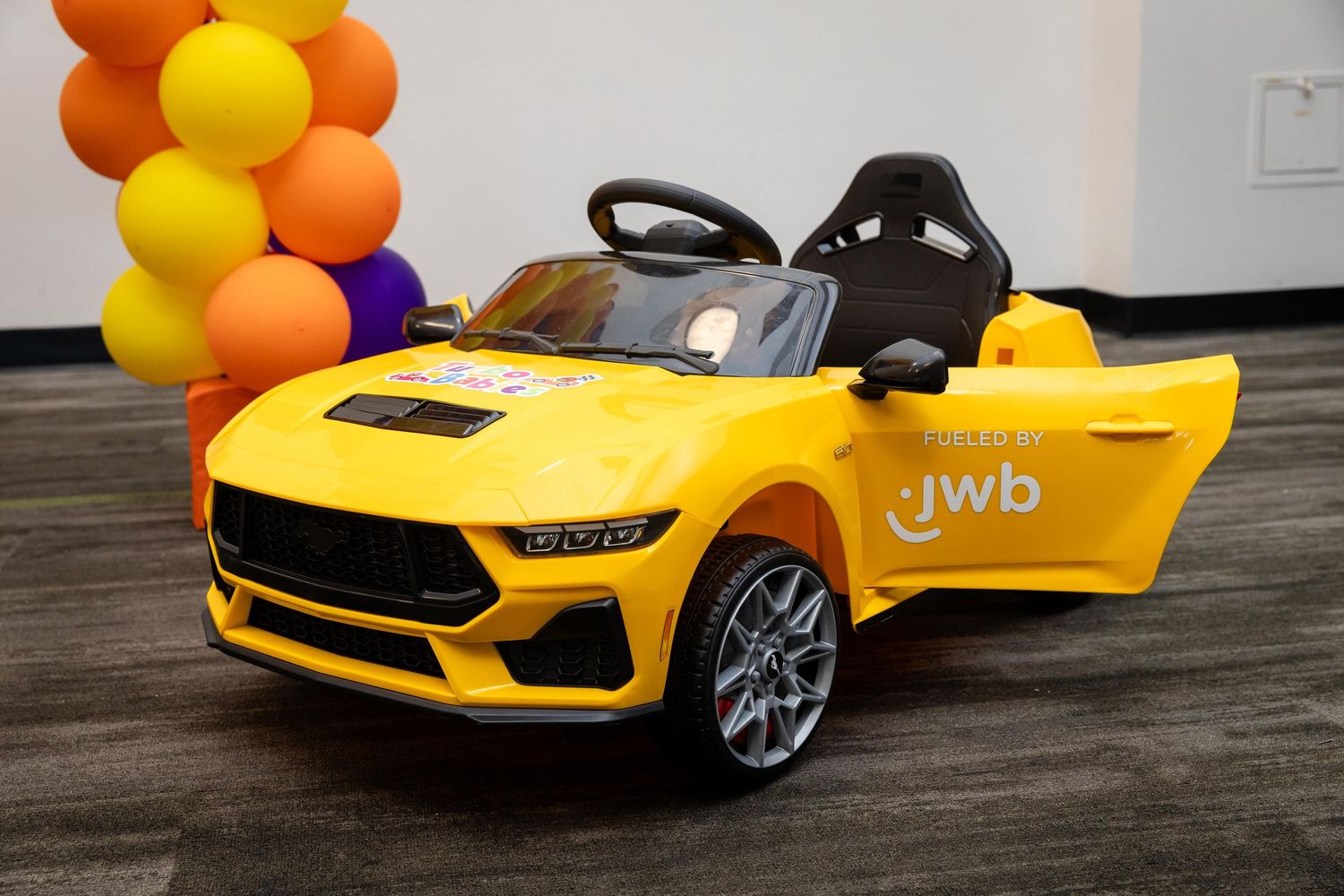 Yellow JWB ride-on toy car for kids displayed indoors with colorful balloon decorations in the background.