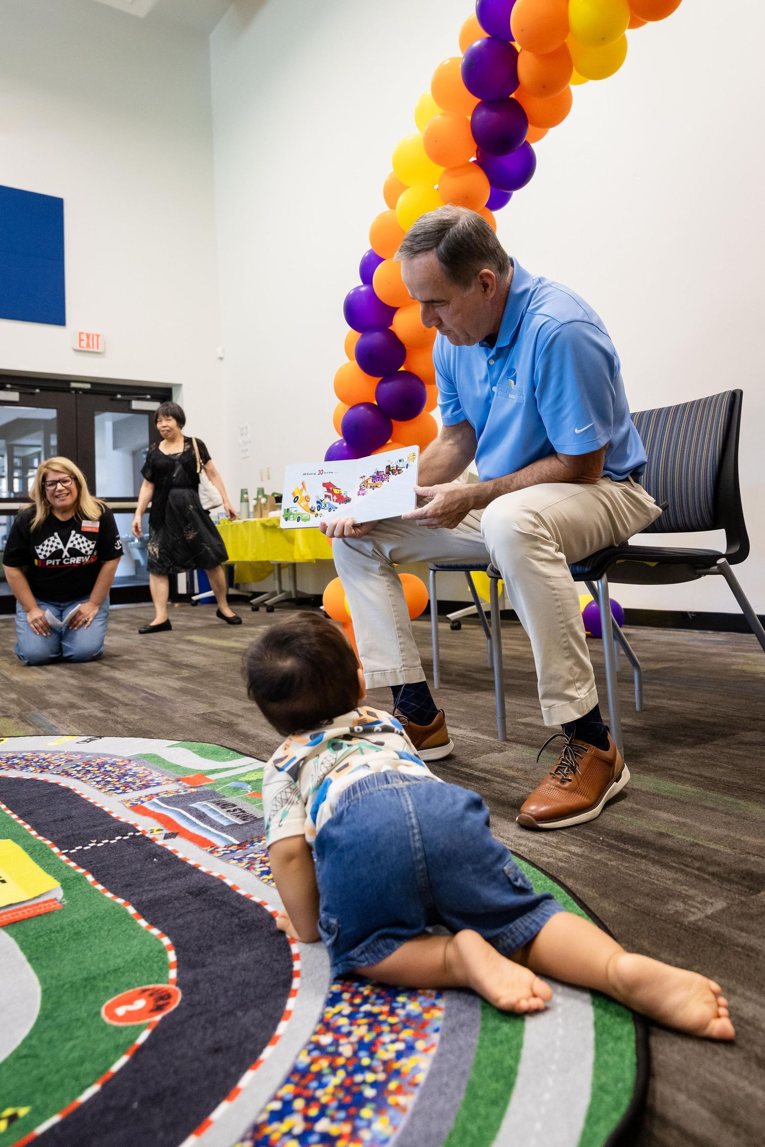 Man in blue shirt reads a colorful book to a toddler on a play mat, with balloon arch decor in background.