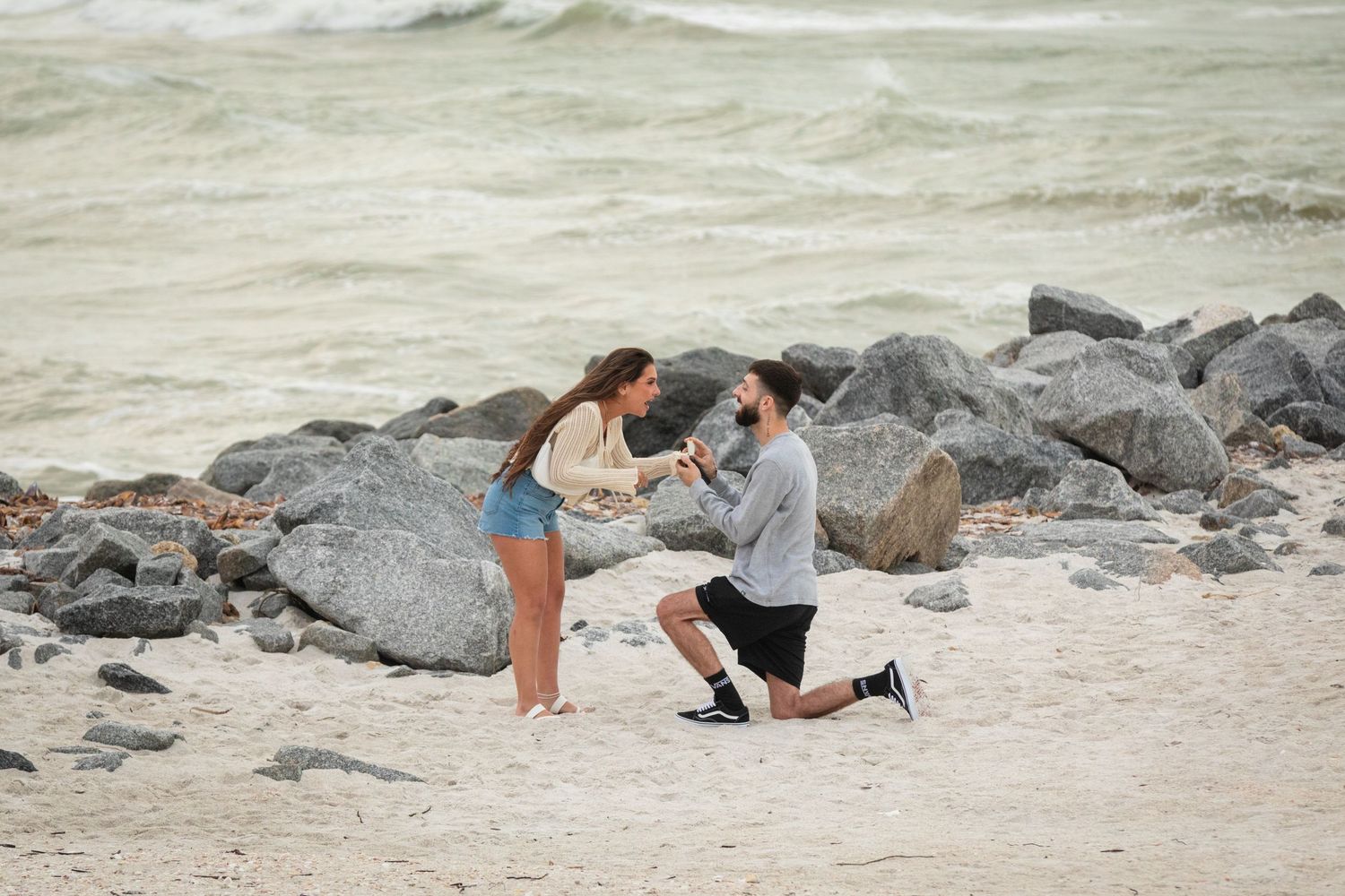 Man proposes to woman on sandy beach with rocky shoreline and ocean waves in the background.