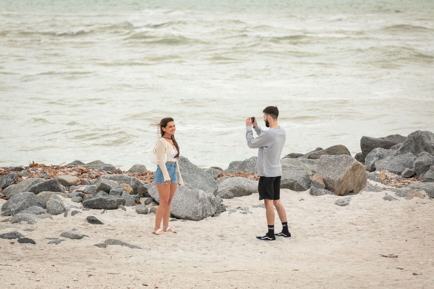 Man photographing woman posing on sandy beach with rocky shoreline and ocean waves in background.