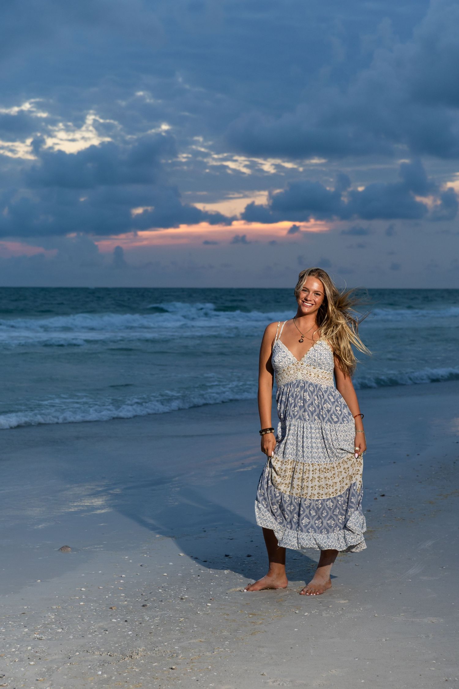 Smiling blonde woman in boho maxi dress walking barefoot on beach at sunset with dramatic cloudy sky.