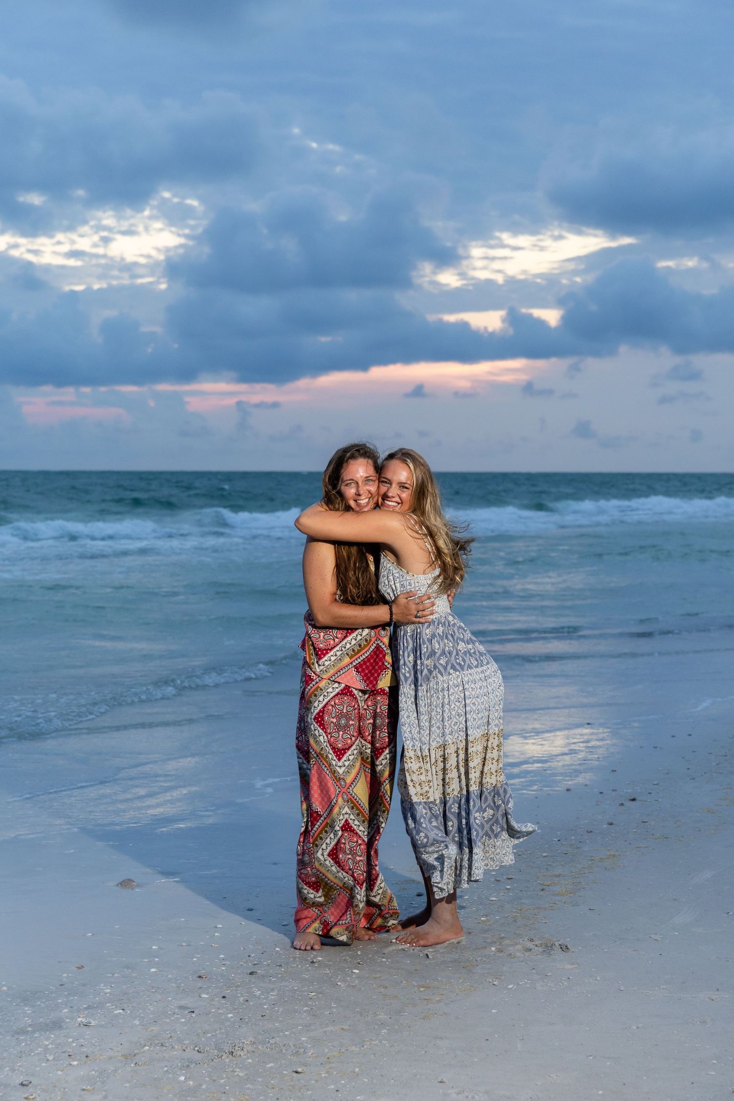 Two women in colorful boho maxi dresses hugging and smiling on a sandy beach at sunset.