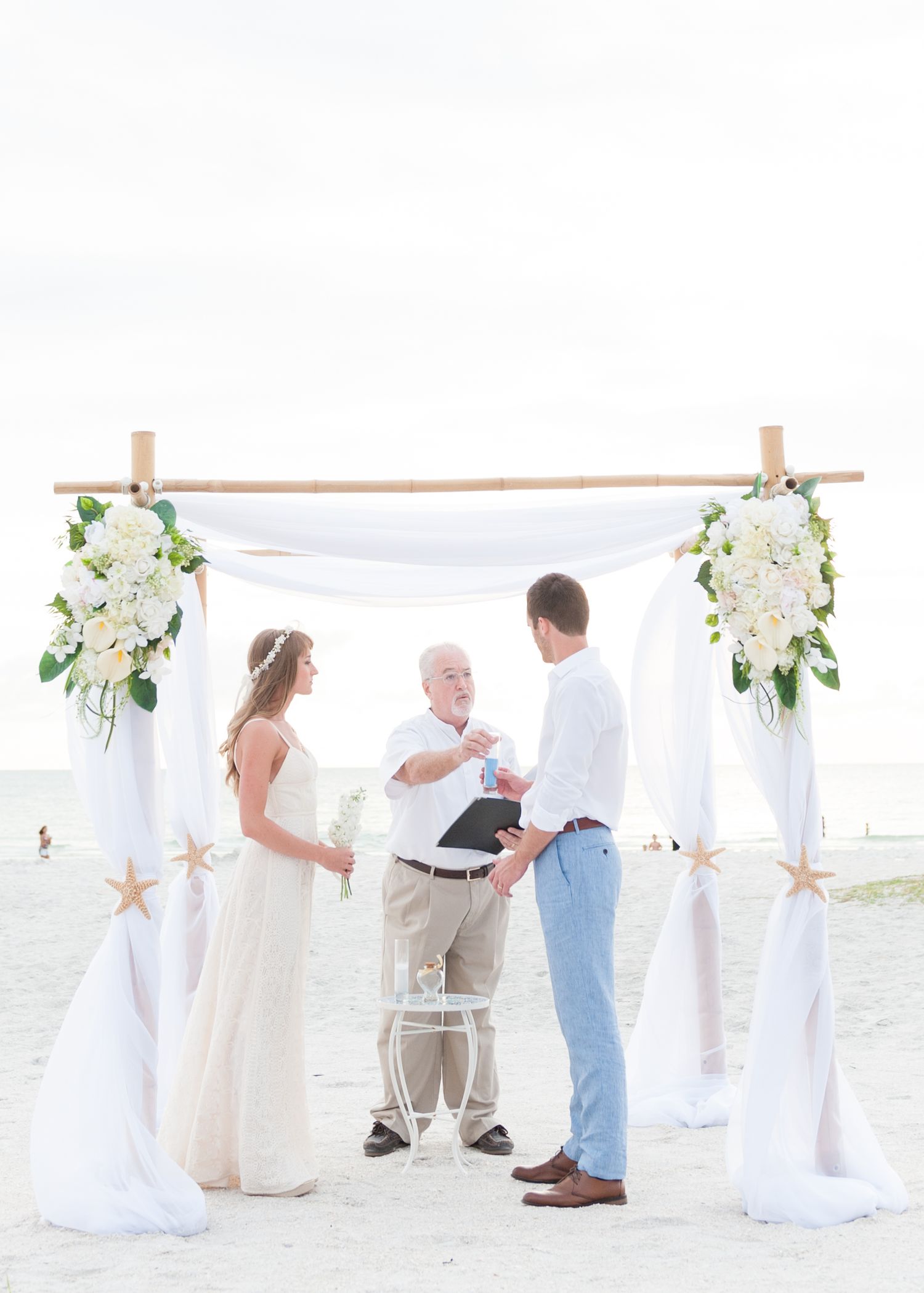 Couple exchanging vows at a beach wedding under a white draped bamboo arch adorned with white flowers and starfish.