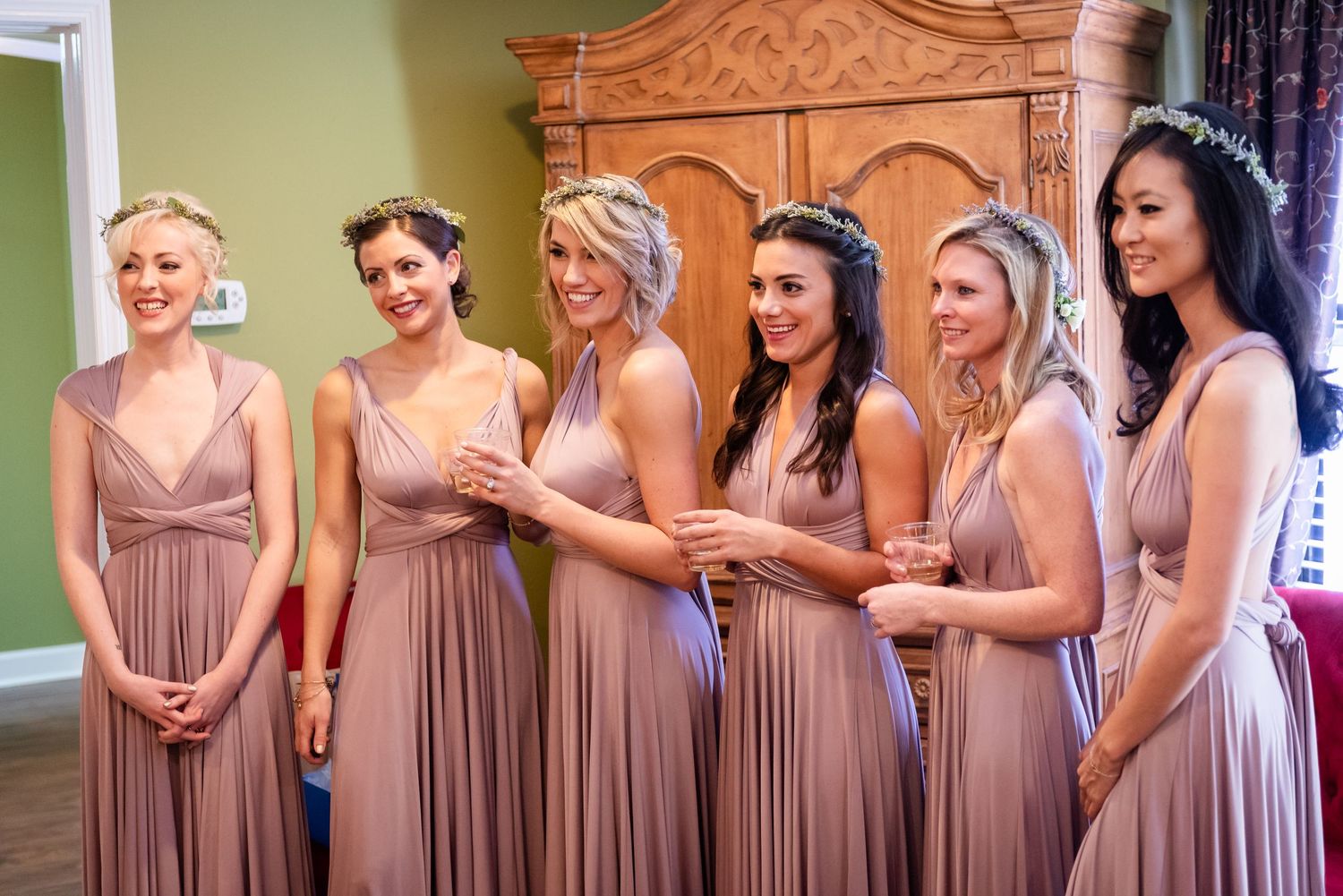 Six bridesmaids in matching dusty rose wrap dresses with floral hair accessories smiling together before a wedding ceremony.