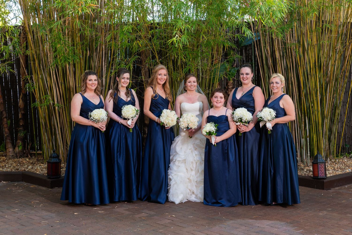 Bride in white gown poses with six bridesmaids in navy blue dresses holding white bouquets before bamboo backdrop.