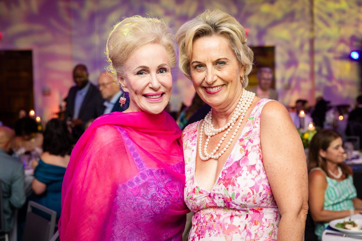 Two elegantly dressed women smiling at a formal gala event, wearing pink outfits and pearl jewelry.