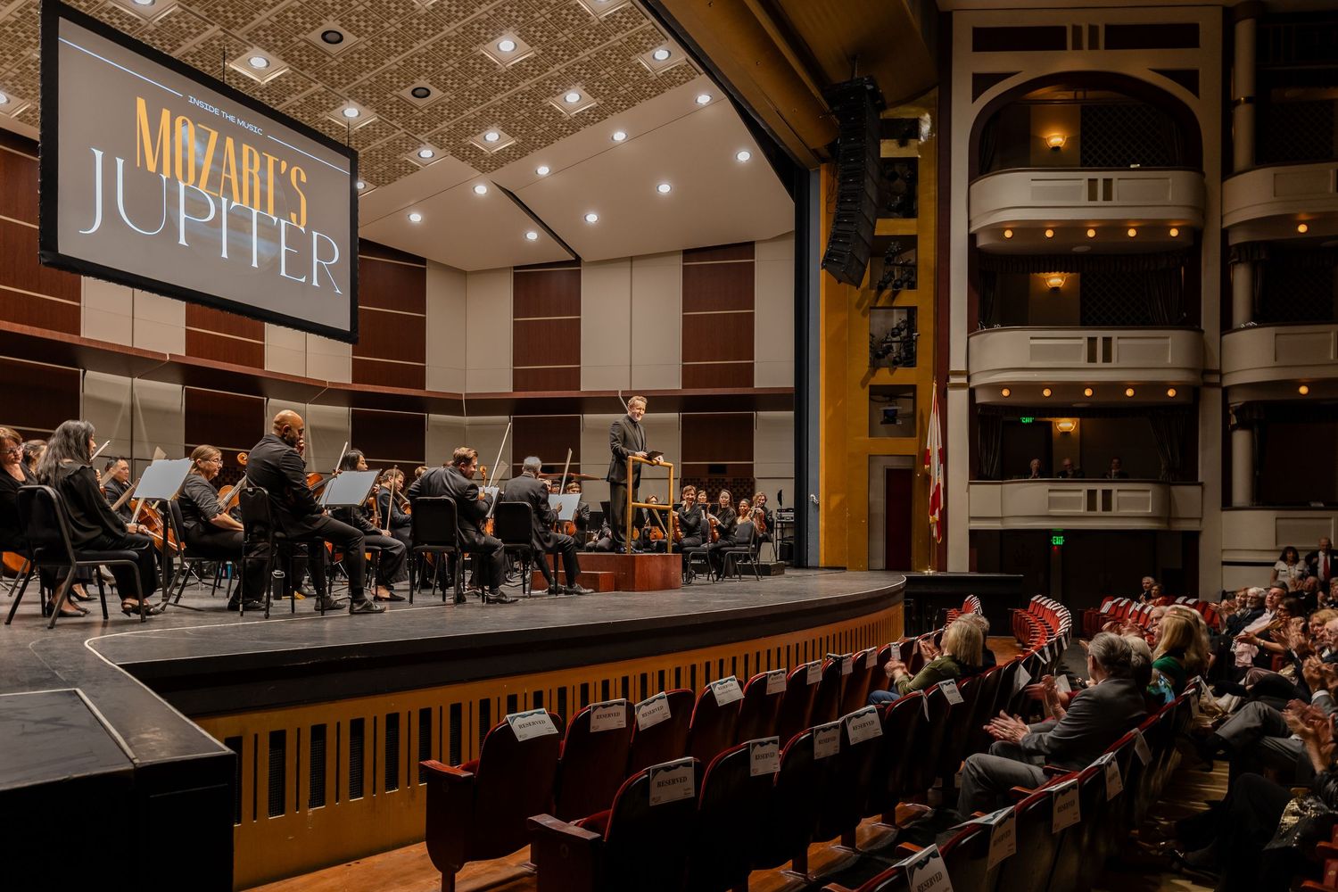 Orchestra performing Mozart's Jupiter on stage in a historic theater with an audience in attendance.