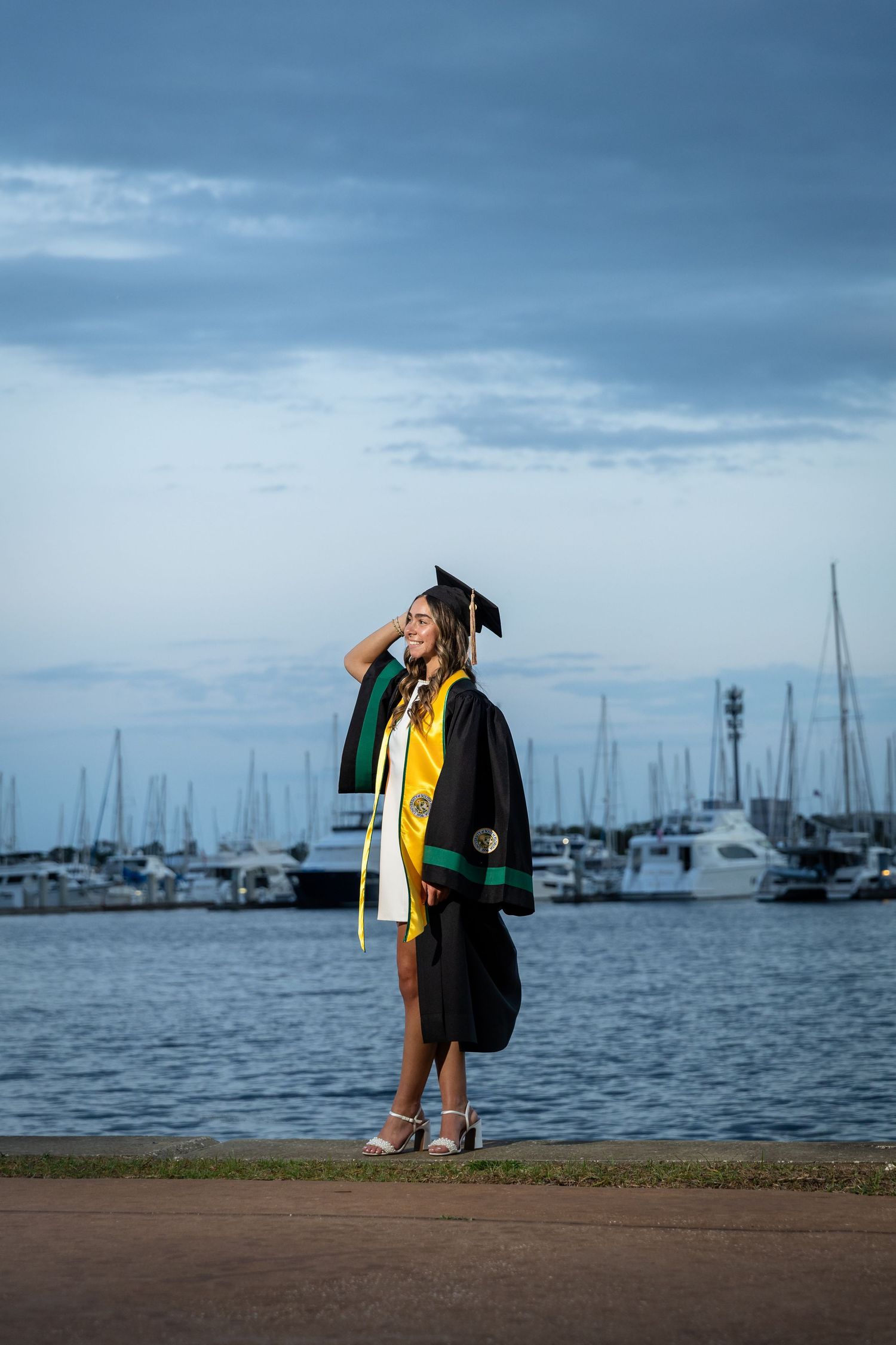 Graduate in black cap and gown with yellow sash poses near marina at dusk.