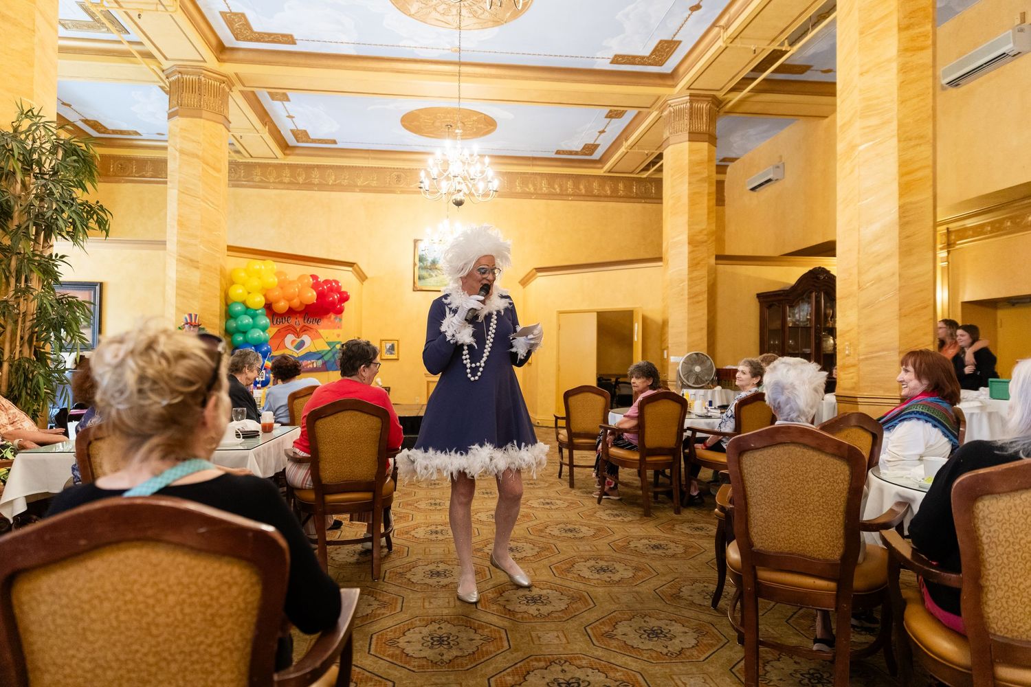 A drag performer in a blue dress entertains seated guests in an elegant gold-decorated dining room.